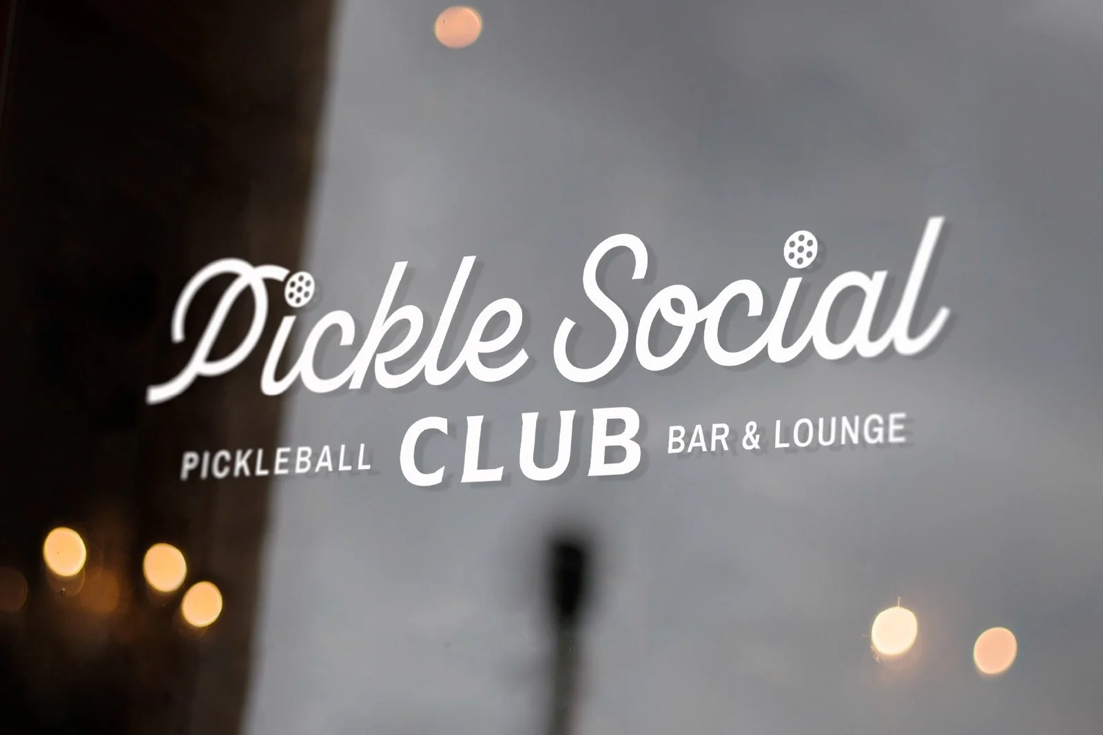 Window sign for Pickle Social, a pickleball club, bar, and lounge, with a blurred background of a cloudy sky and distant lights.