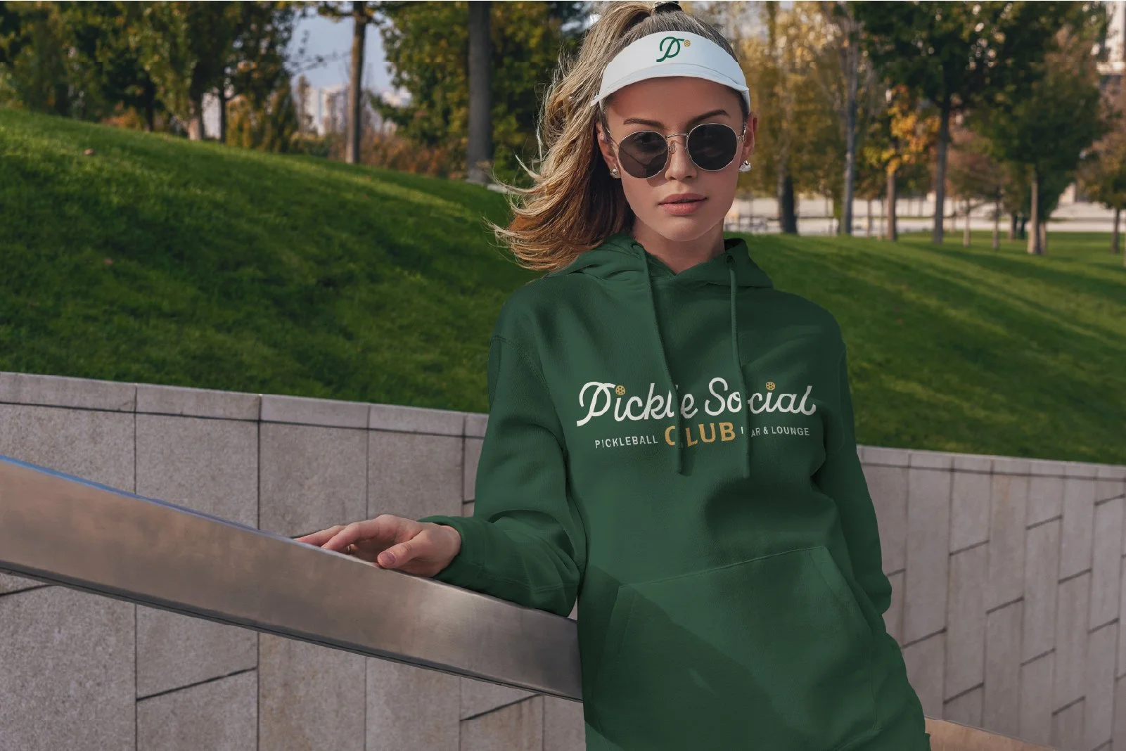 A young woman wearing dark sunglasses, a white visor, and a green hoodie with 'Pickle Social Club' written on it, standing outdoors next to a concrete wall with a grassy park and trees in the background.