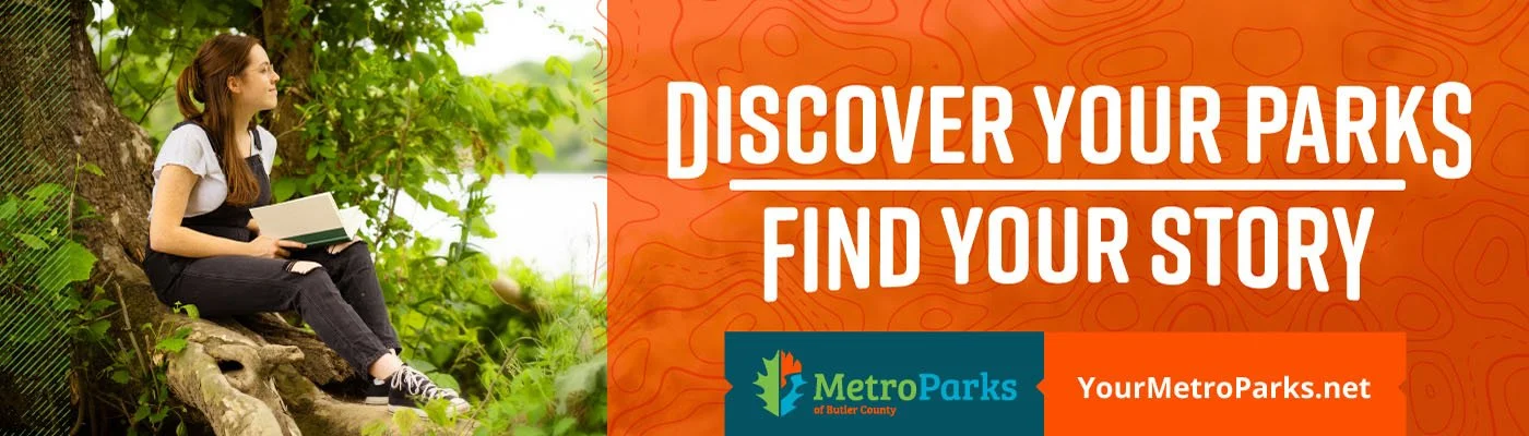 A young woman sitting on a tree root, reading a book beside a river in a park surrounded by green foliage.