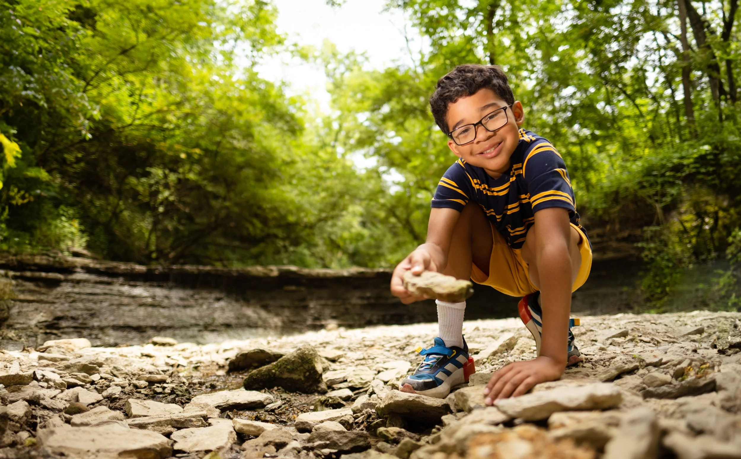 A young boy wearing glasses, a striped shirt, and yellow shorts crouches on a rocky surface in a forest, smiling and holding a rock.