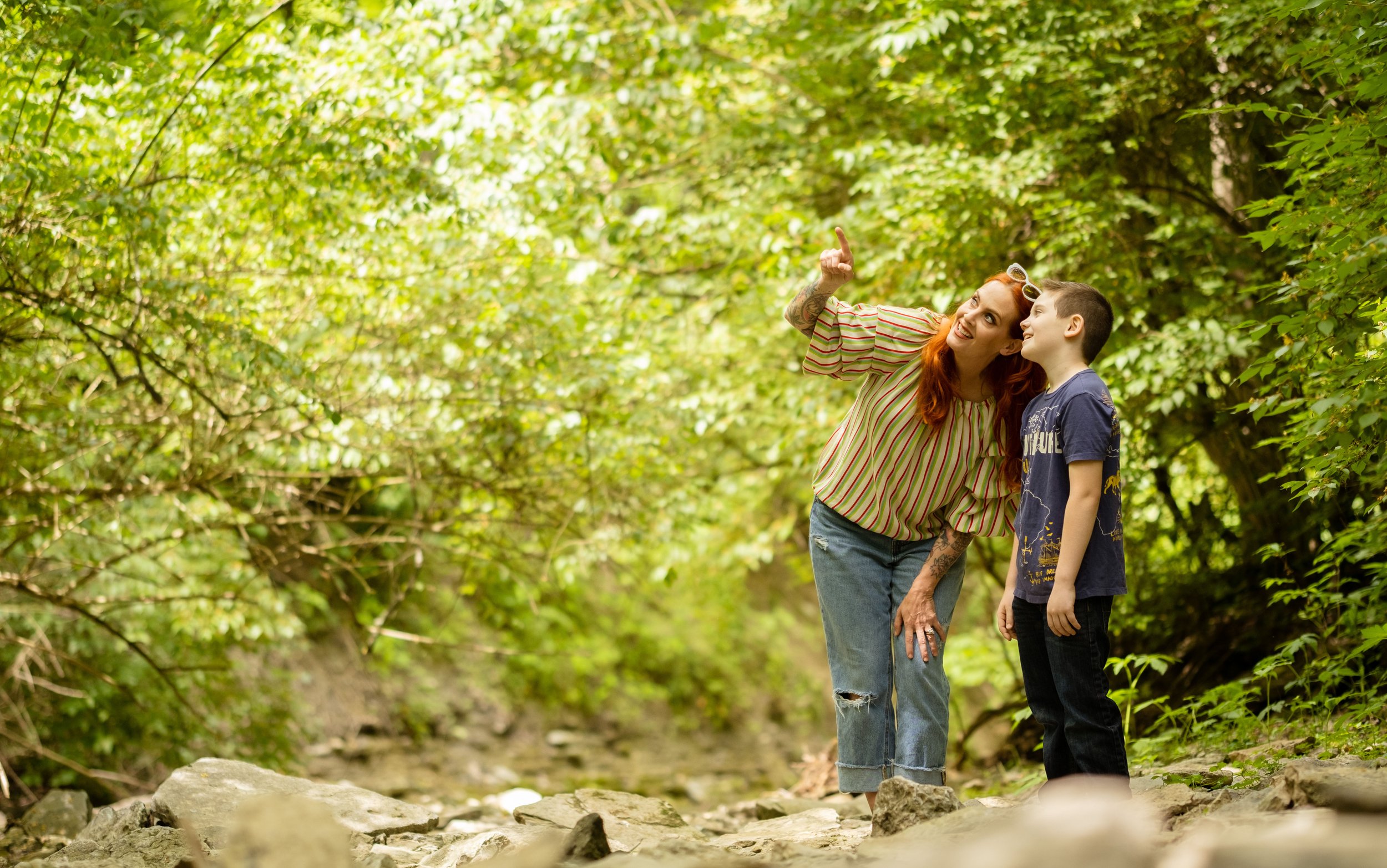 A woman with red hair and tattoos pointing and talking to a young boy in a wooded area with green leaves.