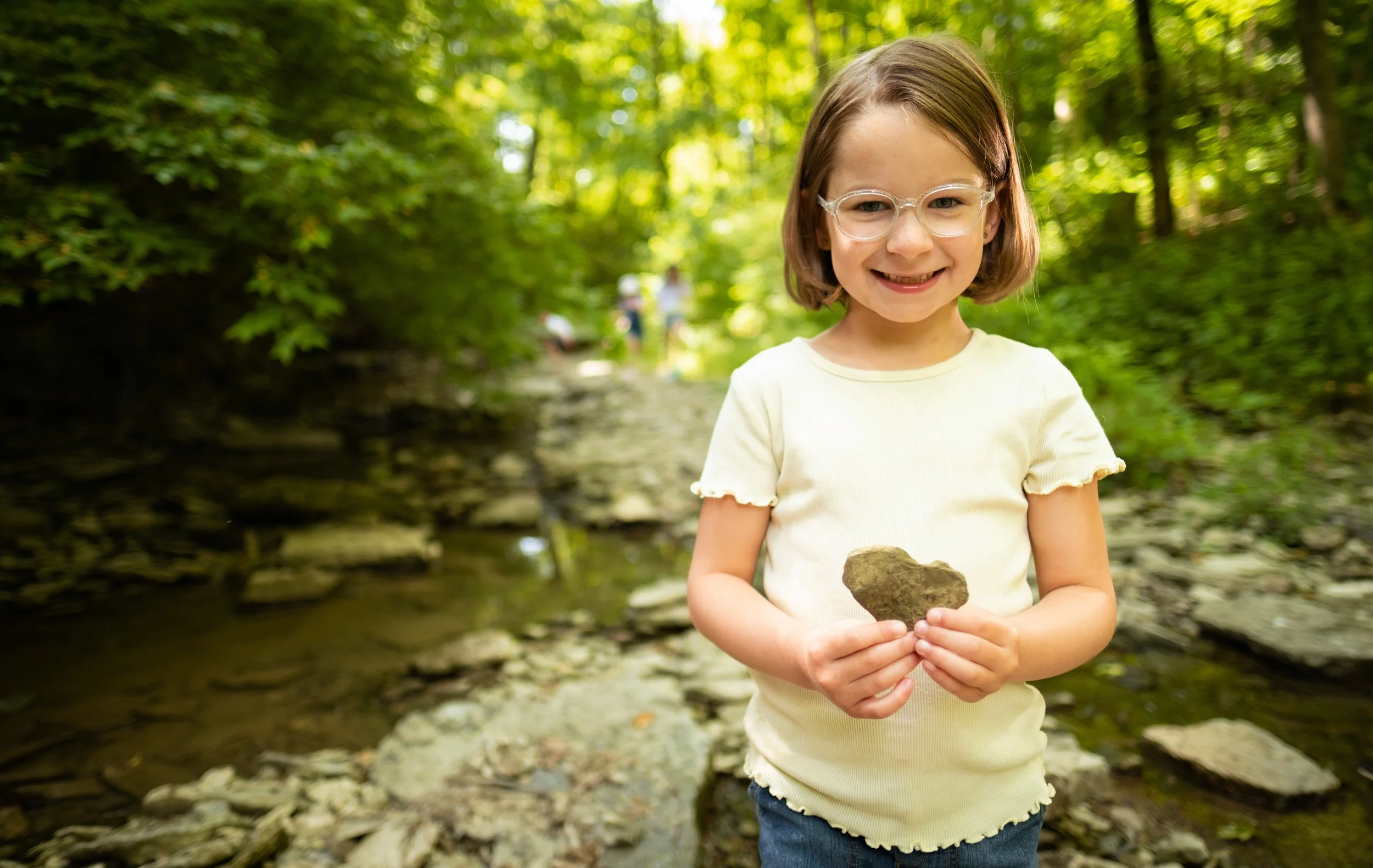 A young girl with glasses smiling and holding a rock in her hands outdoors in a wooded area.