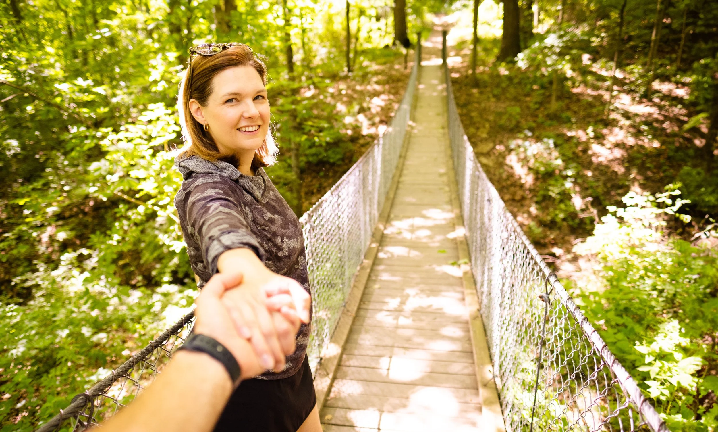 A woman smiling and holding hands with another person on a suspension bridge in a lush green forest.