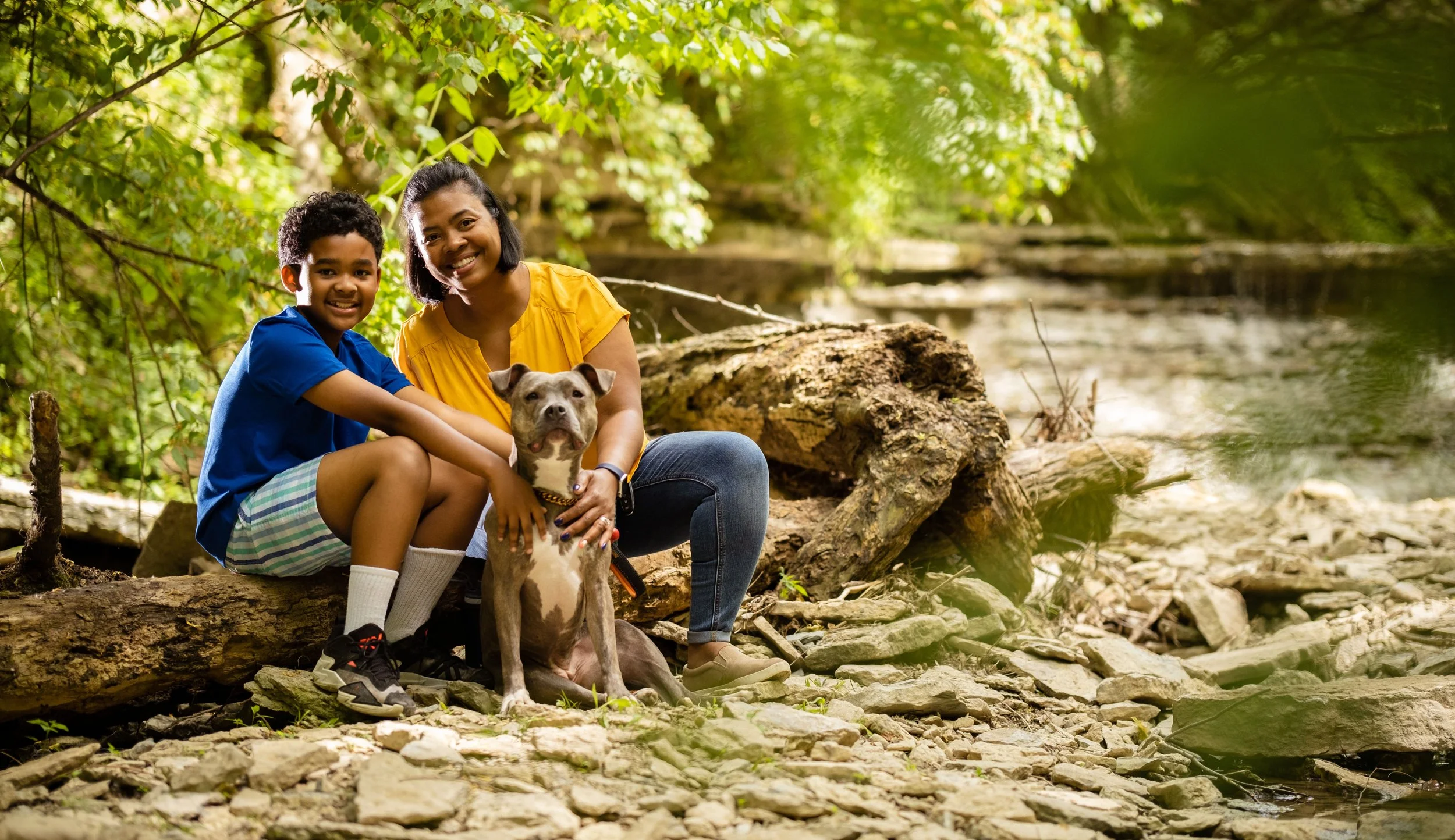 A woman and a boy sitting on a log by a creek in a forest, smiling, with a dog sitting in front of them.