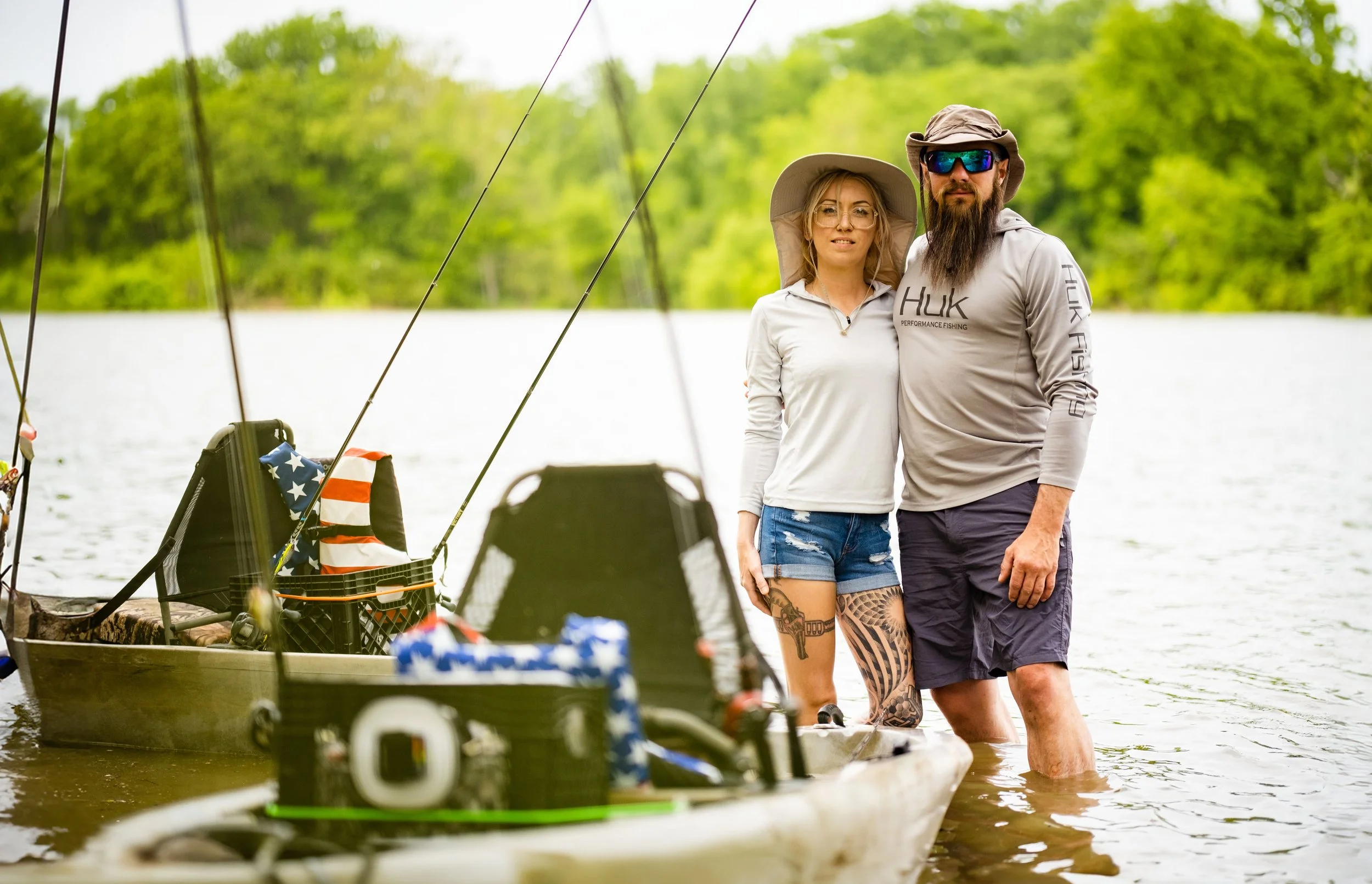 A man and woman standing knee-deep in a lake, posing with a boat and fishing gear. The woman is wearing a wide-brimmed hat and sunglasses, while the man is wearing a wide-brimmed hat and sunglasses. The background shows lush green trees.
