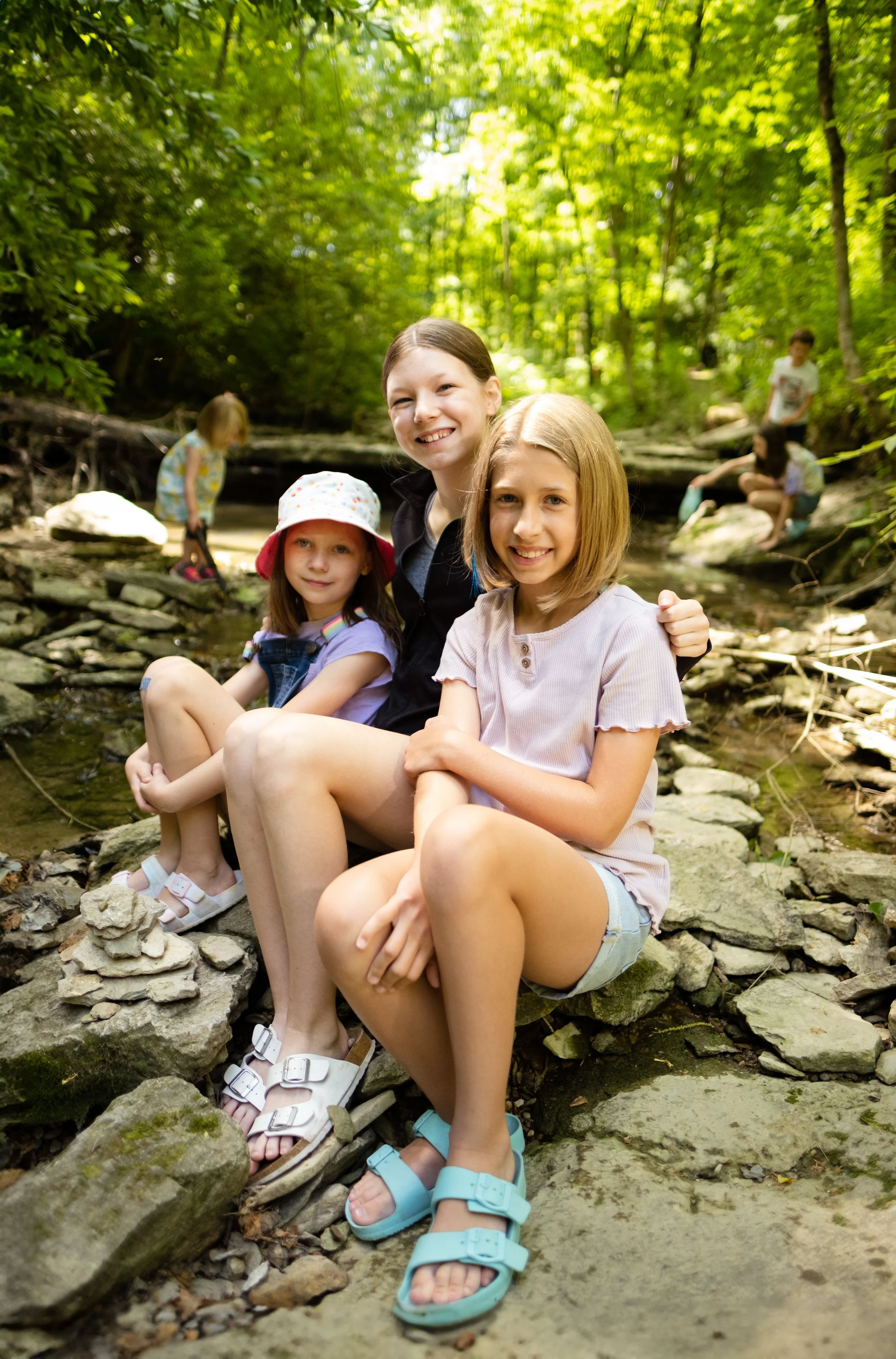 A group of children sitting on rocks in a creek surrounded by green trees, enjoying a day outdoors.
