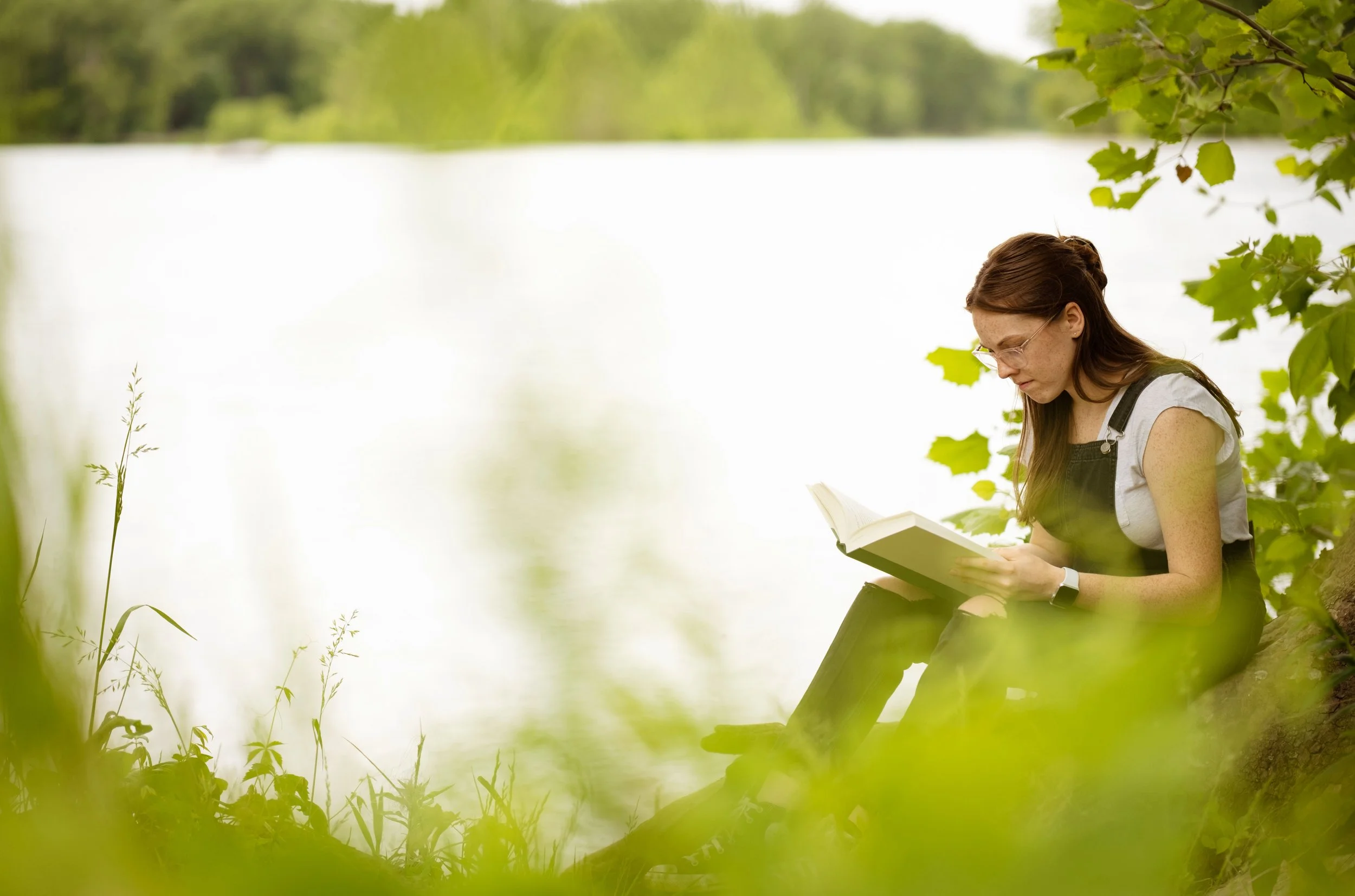 A woman sitting on the grass by a lake, reading a book, surrounded by green leaves and trees.