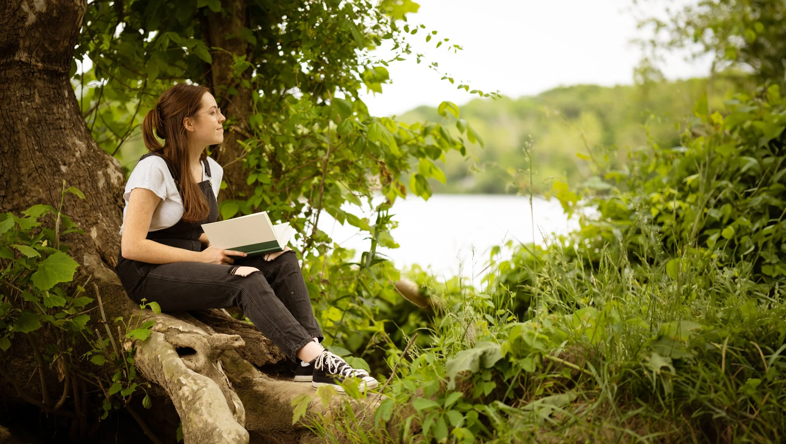 A young woman sitting on a tree root near a lake, reading a book in a lush, green outdoor setting.