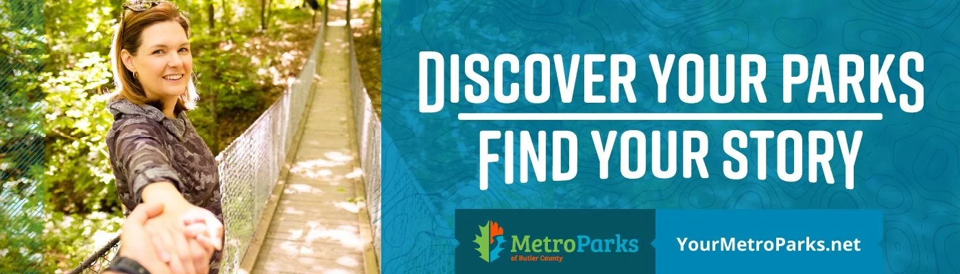A smiling woman holding hands while walking on a suspension bridge in a park surrounded by green trees, with a graphic overlay promoting Metro Parks of Butler County.
