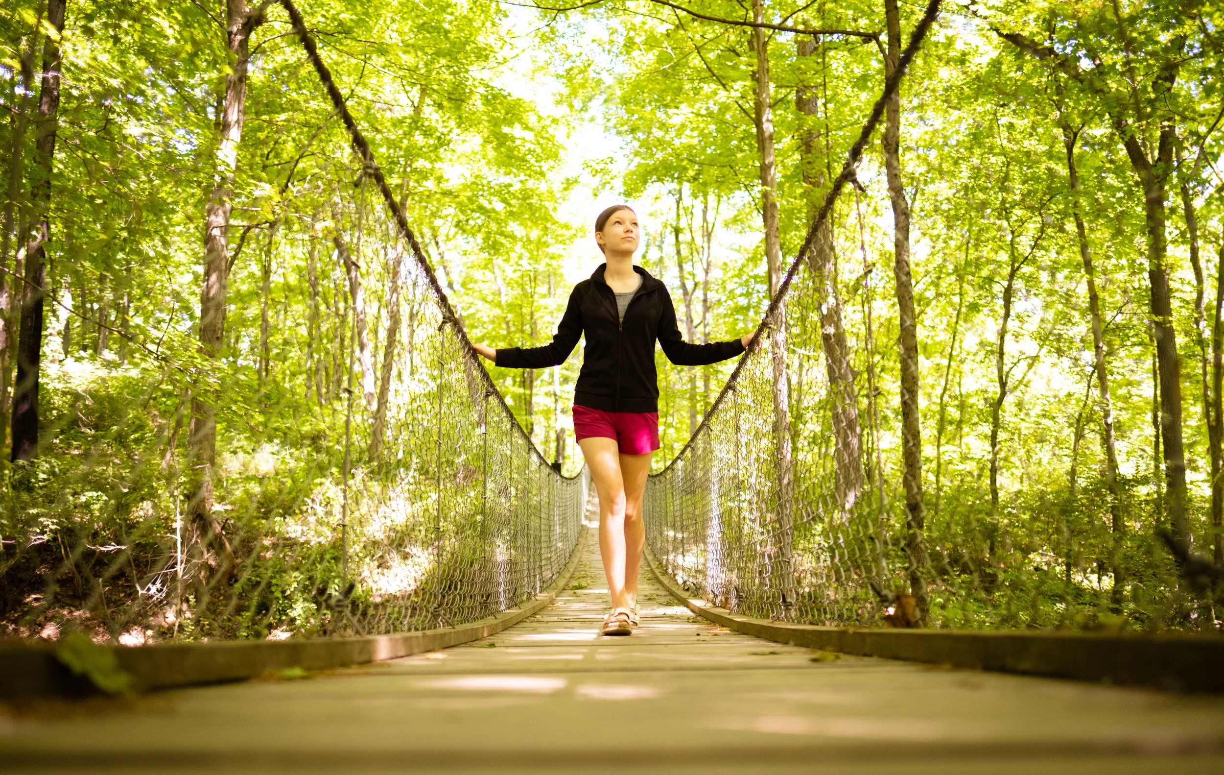 A woman walking on a narrow suspension bridge in a lush green forest during daytime.