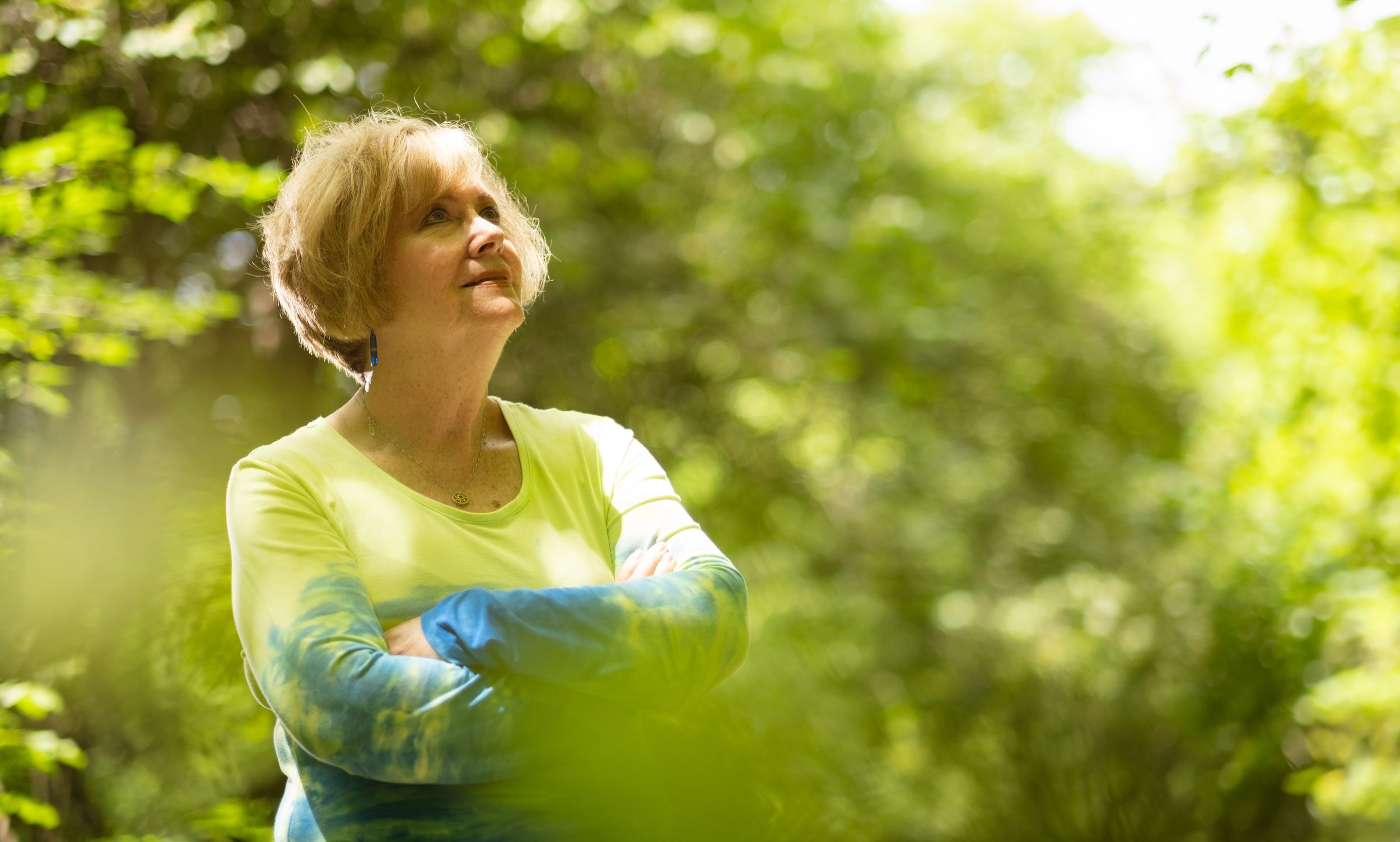 A woman with blonde hair, wearing a yellow and blue tie-dye long sleeve shirt, is standing outdoors in a green, leafy forest with her arms crossed, looking upwards.