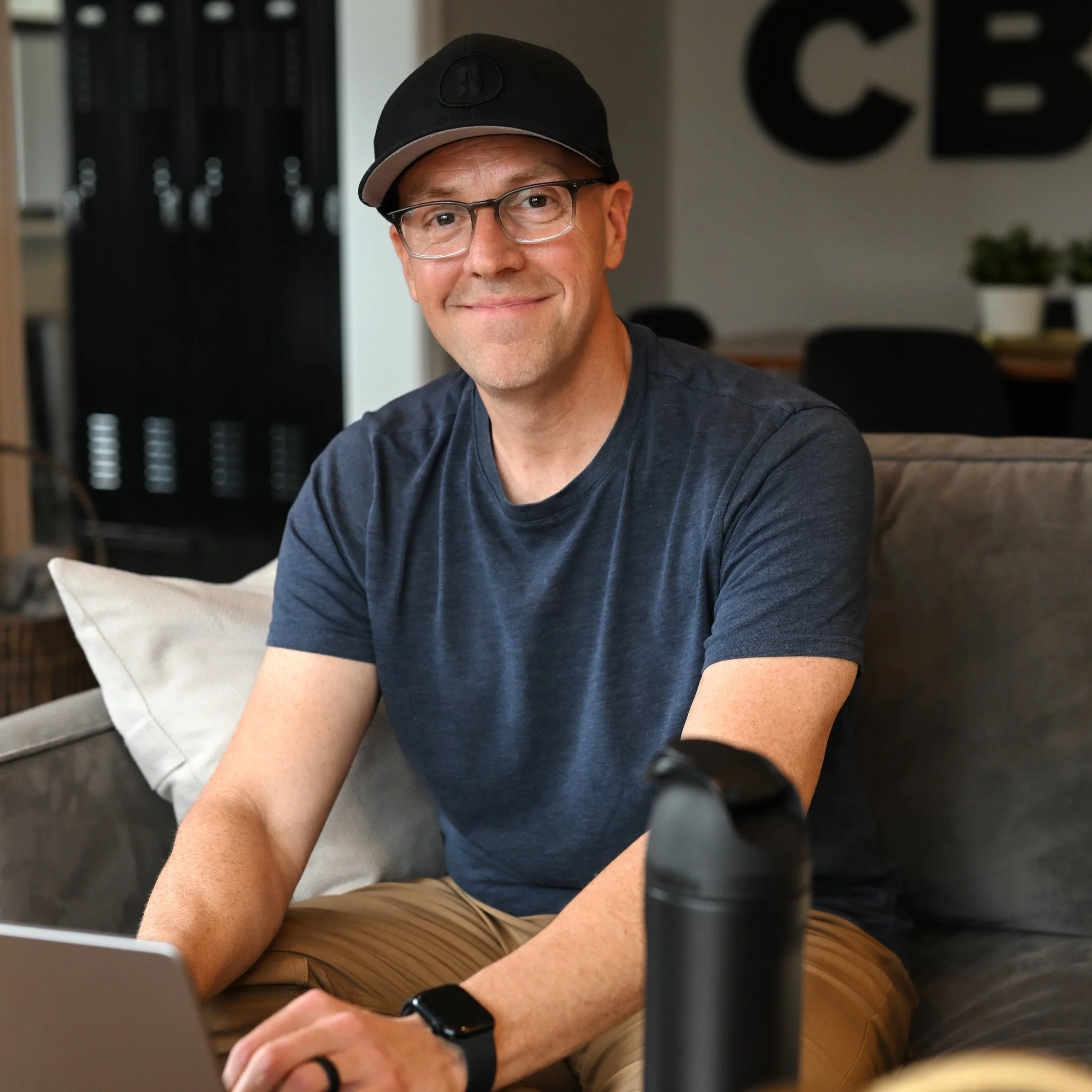 A man wearing glasses, a black cap, and a blue t-shirt sitting on a couch in a modern room.