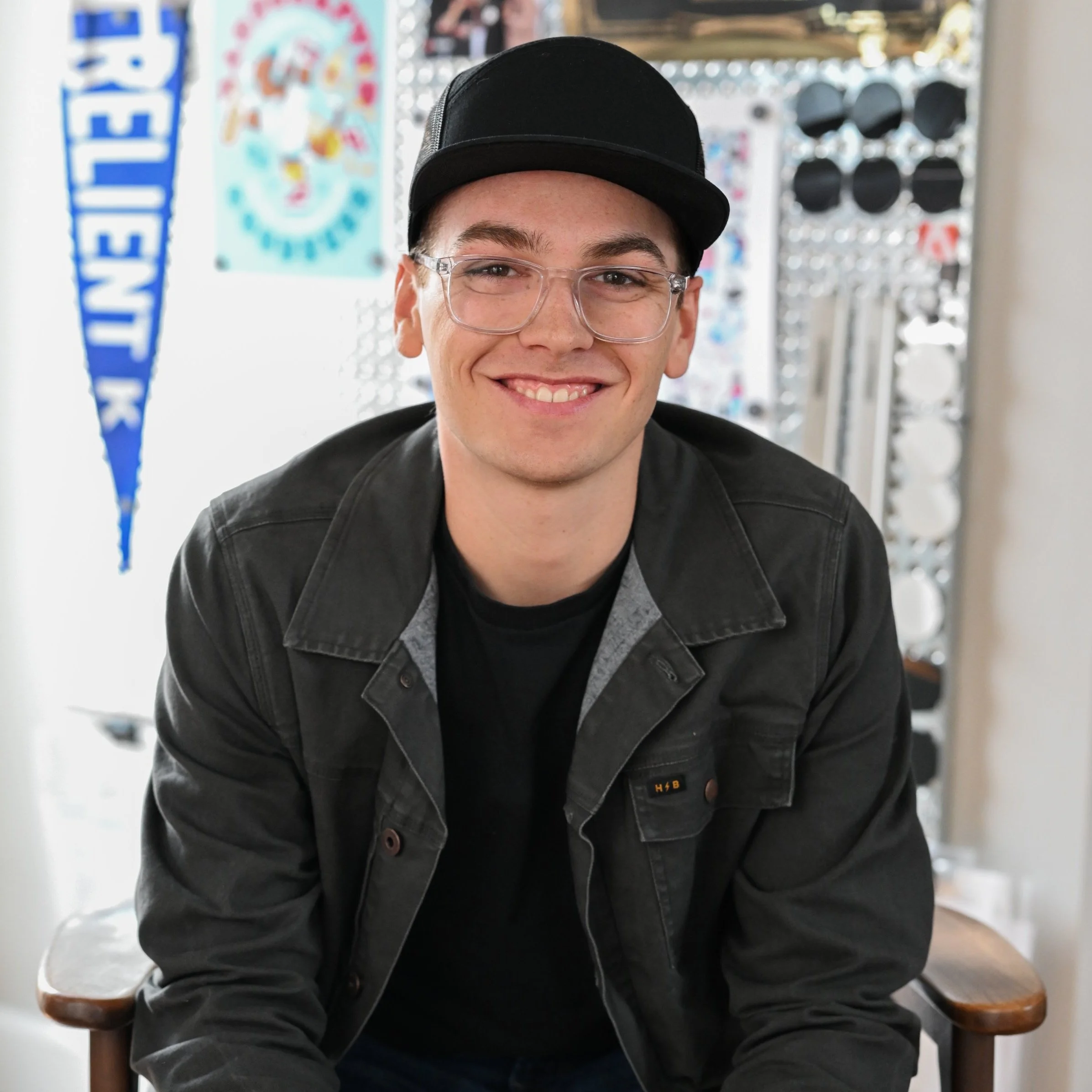 A young man with glasses and a black baseball cap smiling, sitting on a chair indoors with a blue and white pennant and colorful artwork in the background.