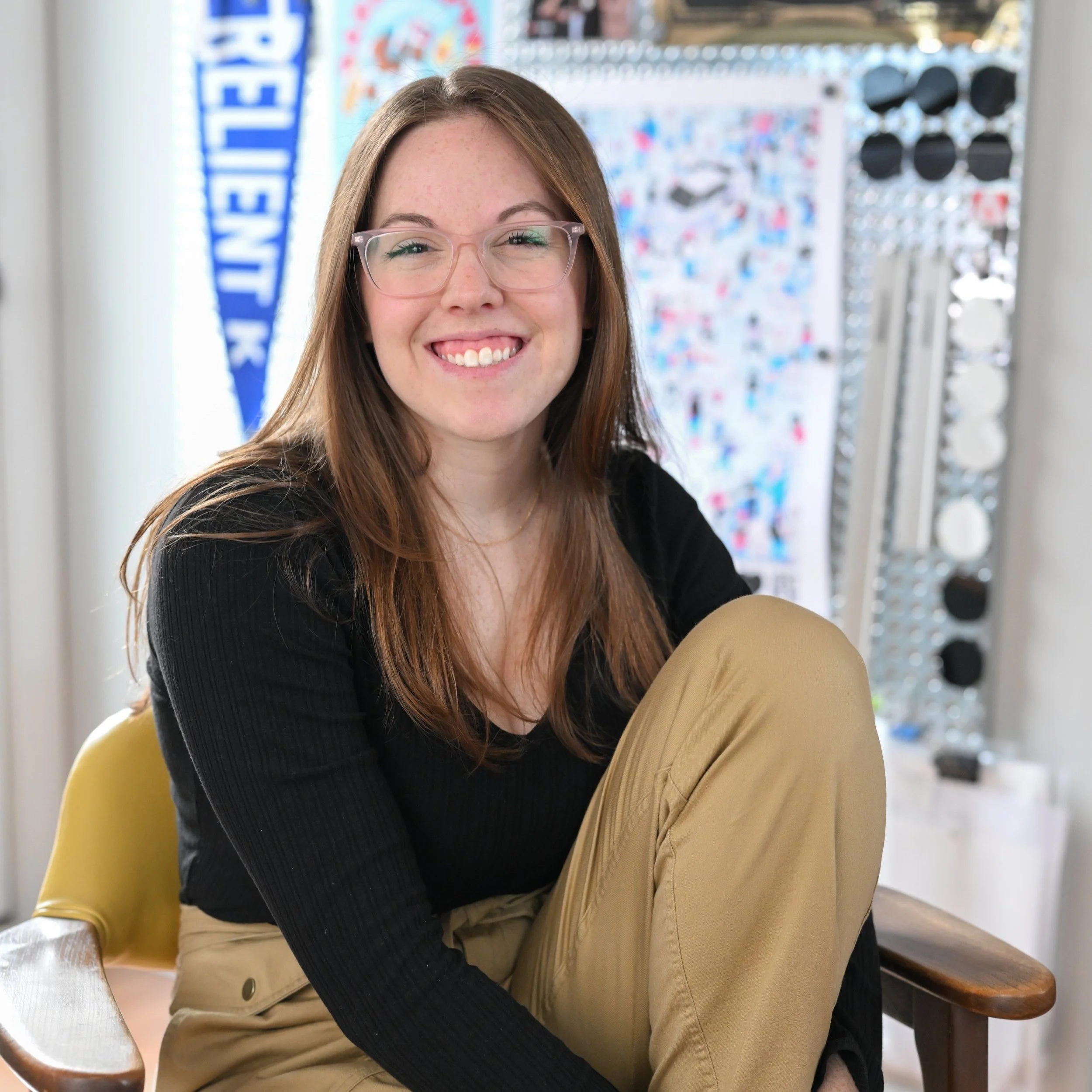 A young woman with long reddish-brown hair, glasses, and a black shirt smiling while seated on a yellow chair in a room with a colorful background, including a mirror and a blue and white Penn State banner.