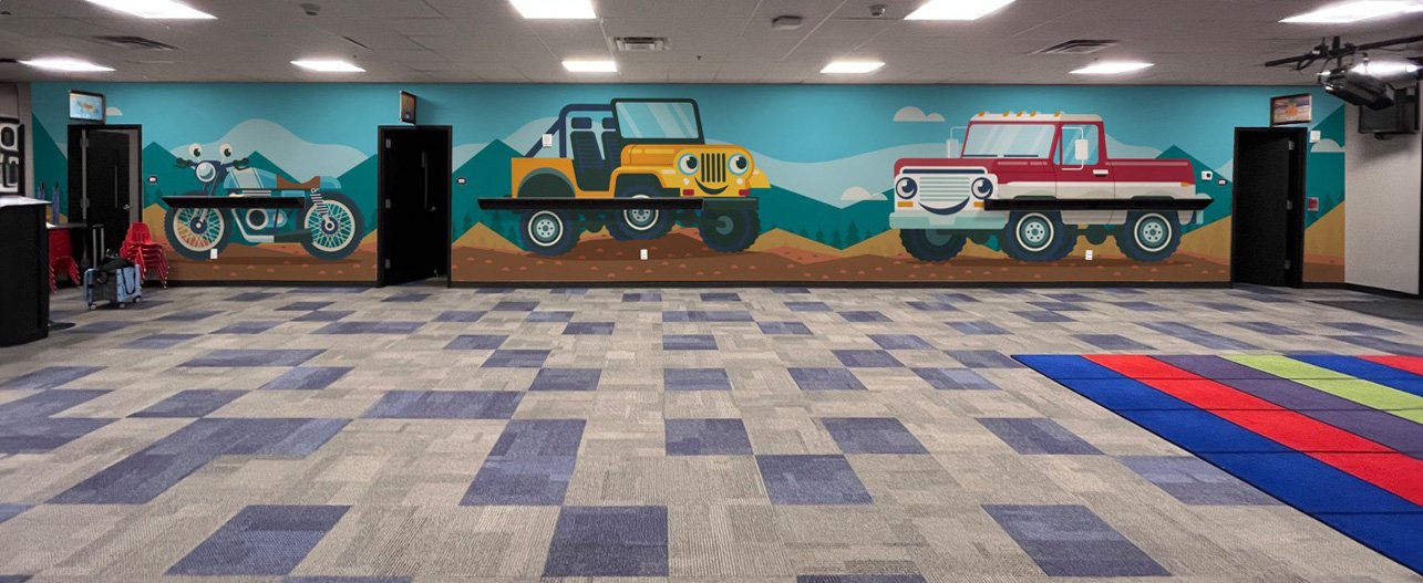 Colorful mural of off-road vehicles, including a motorcycle, a yellow jeep, and a red truck, painted on a light blue wall in an indoor play area with a patterned carpet and a multicolored mat.