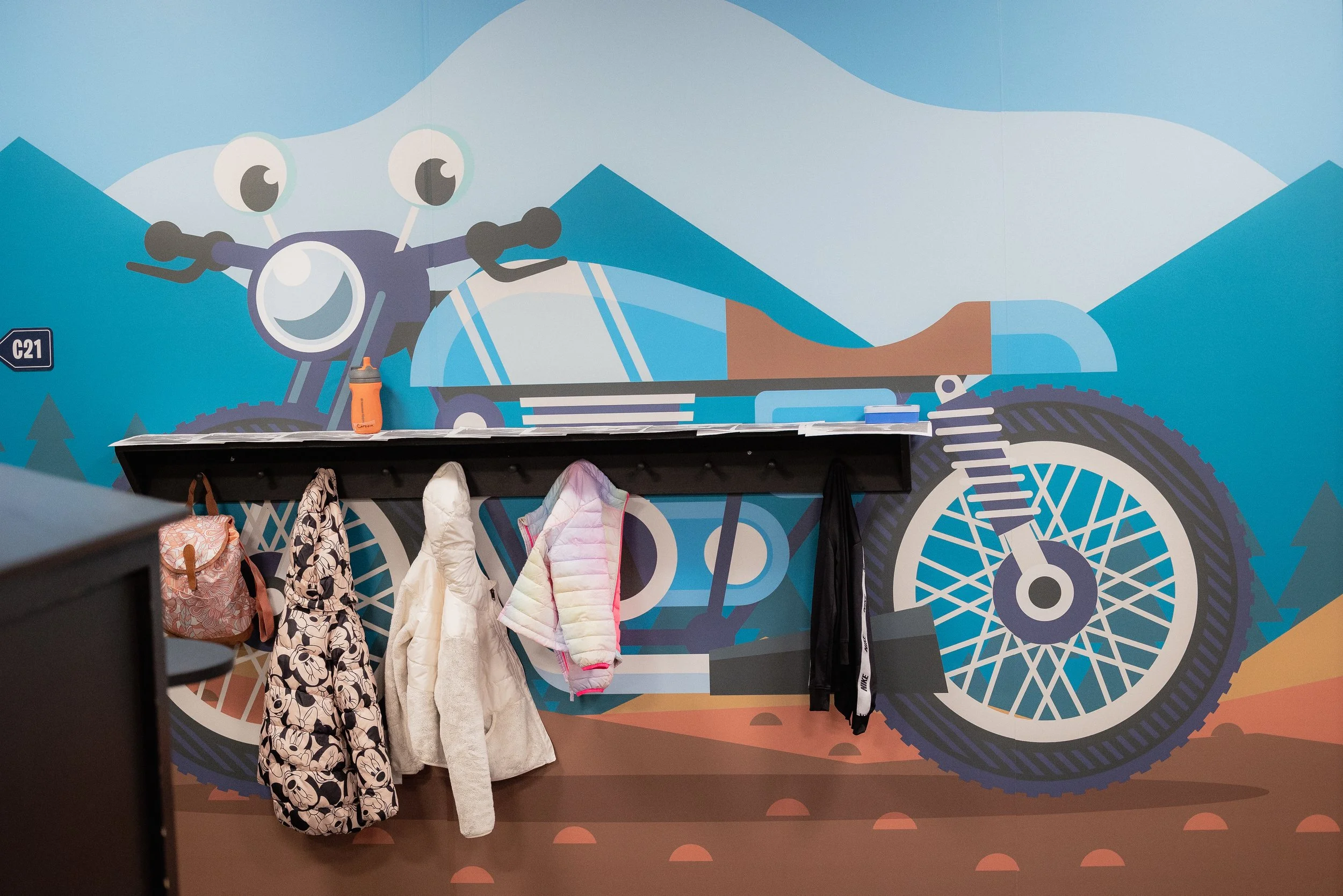 Colorful mural of a motorcycle with mountains in the background, with hanging jackets and backpacks on a black shelf in front of it.