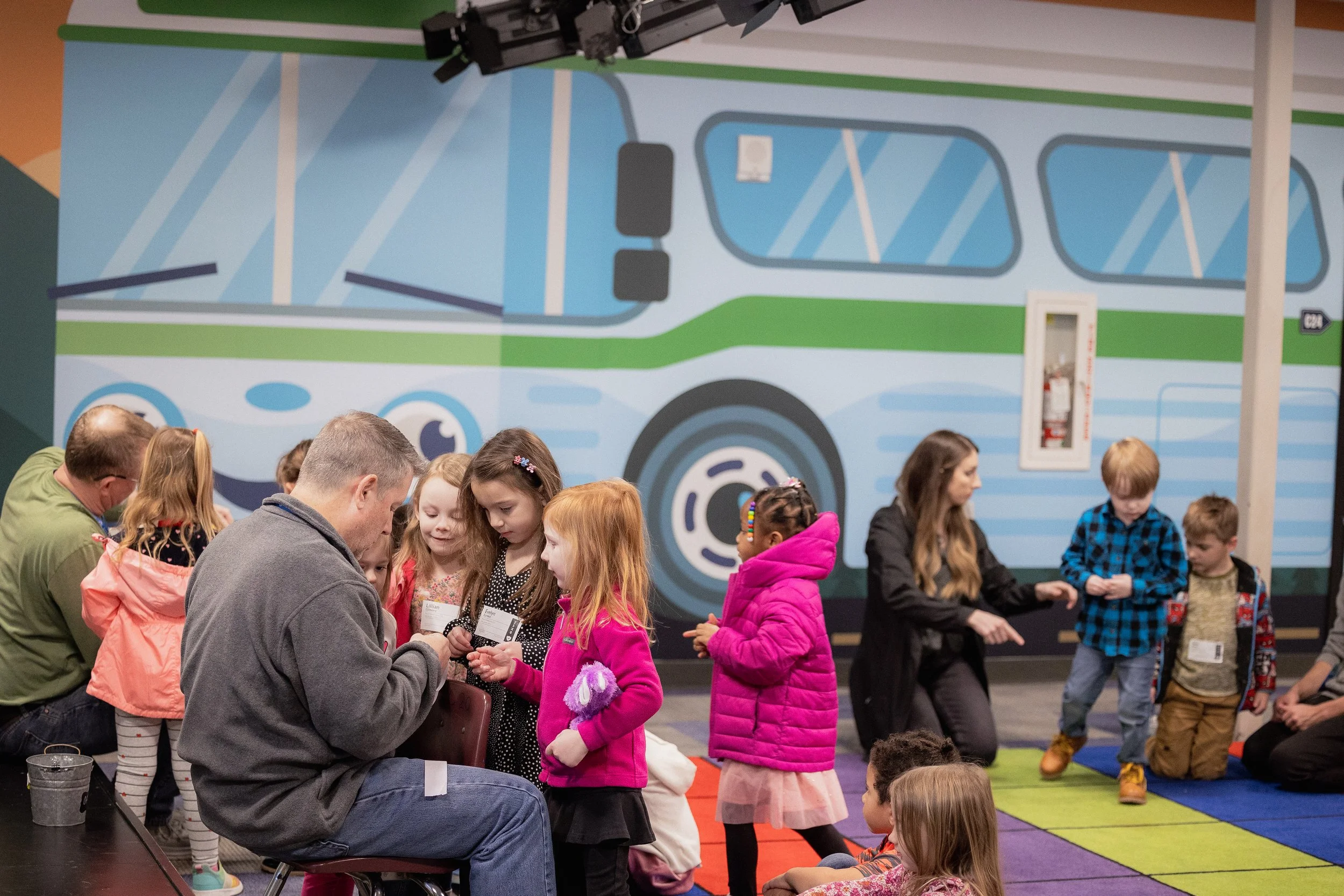 Children and adults gathered indoors in front of a large colorful bus mural.