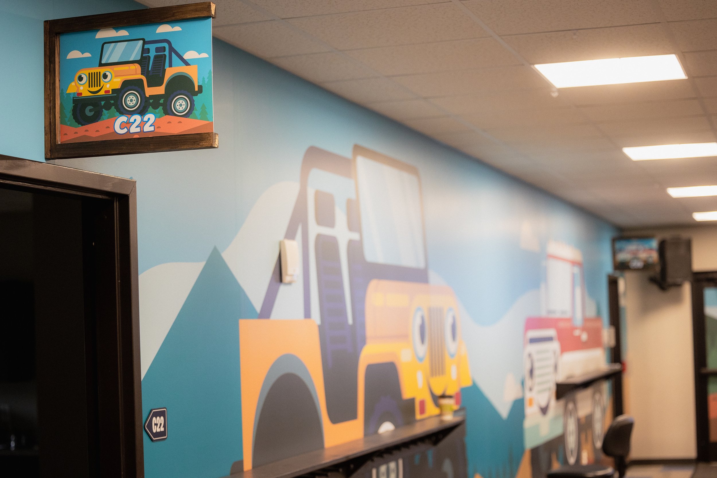 Wall mural featuring cartoon construction vehicles, including a yellow bulldozer with big eyes and a smiling face, painted on a light blue background with mountains and clouds.