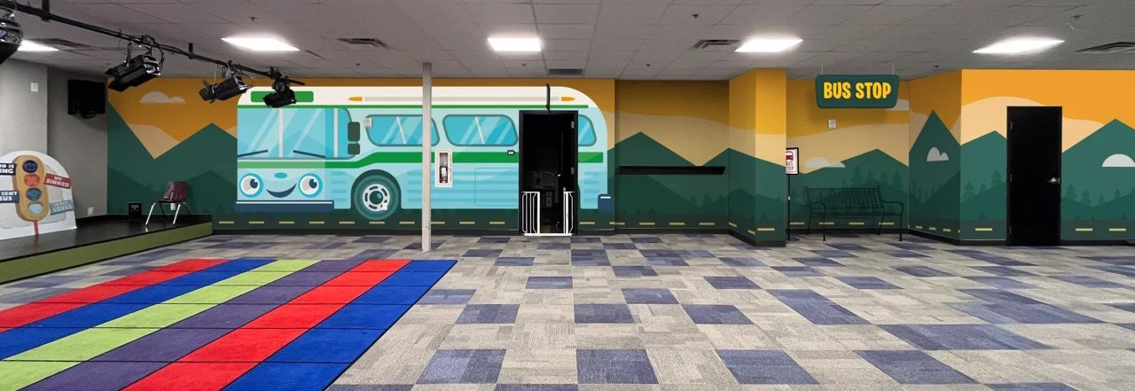 Indoor children's play area with a colorful striped carpet, a wall mural of a bus with mountain scenery, benches, and a bus stop sign.