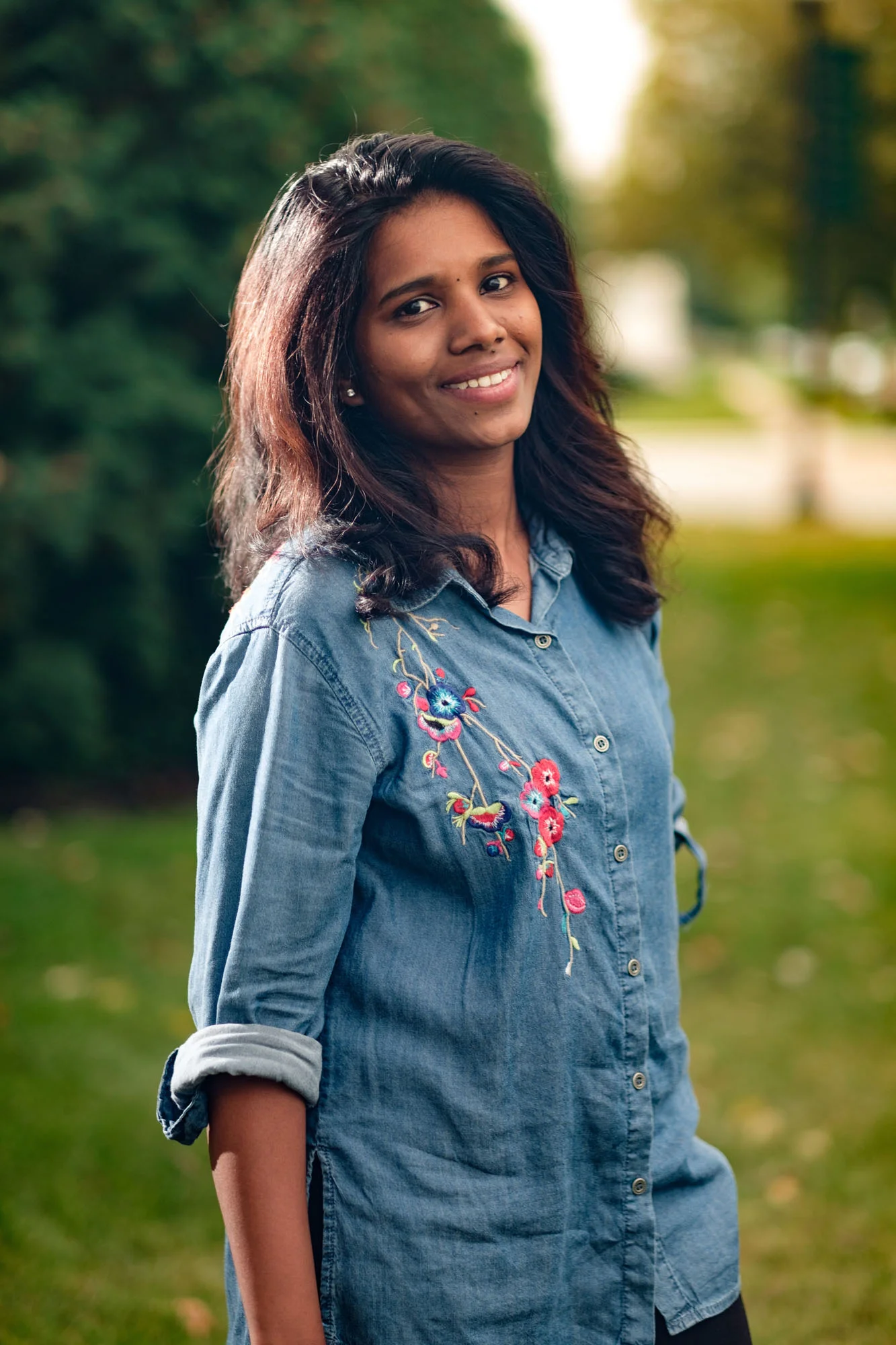 A young woman with dark hair and a gentle smile, standing outdoors in a park with lush green trees and grass, wearing a denim shirt with floral embroidery on the shoulder.