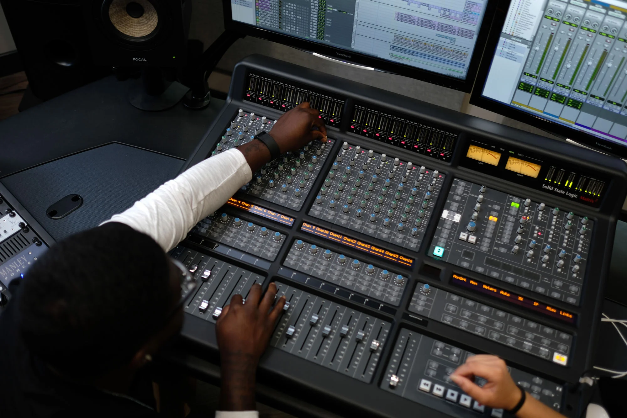 A person operating a professional audio mixing console with two computer screens displaying digital audio workstation software.