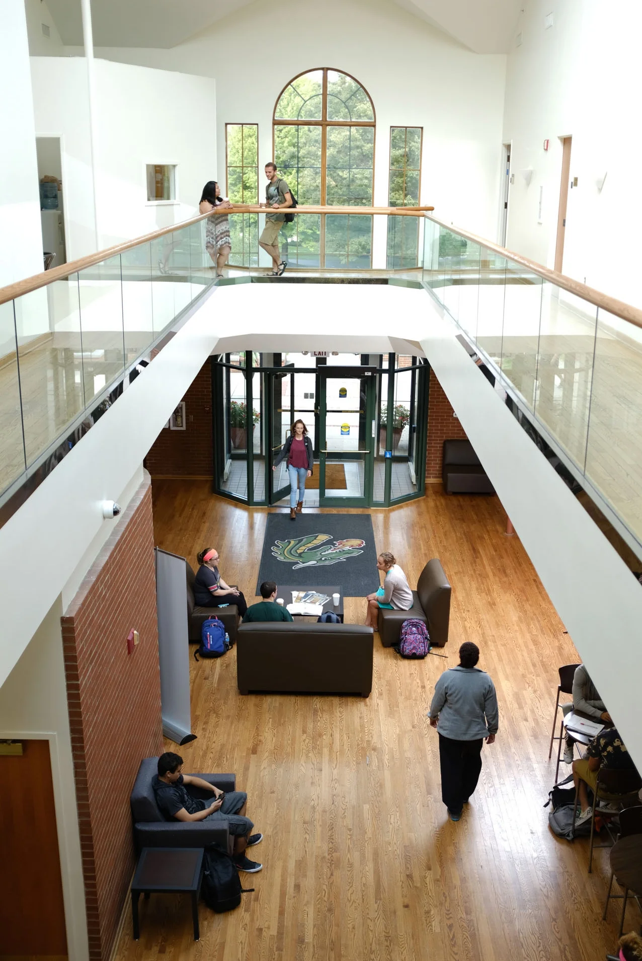 A spacious indoor university or college lobby with a two-story design, featuring a wooden floor, brick accents, and a large arched window. Students and visitors are sitting and walking, with some near the entrance and others lounging on couches.