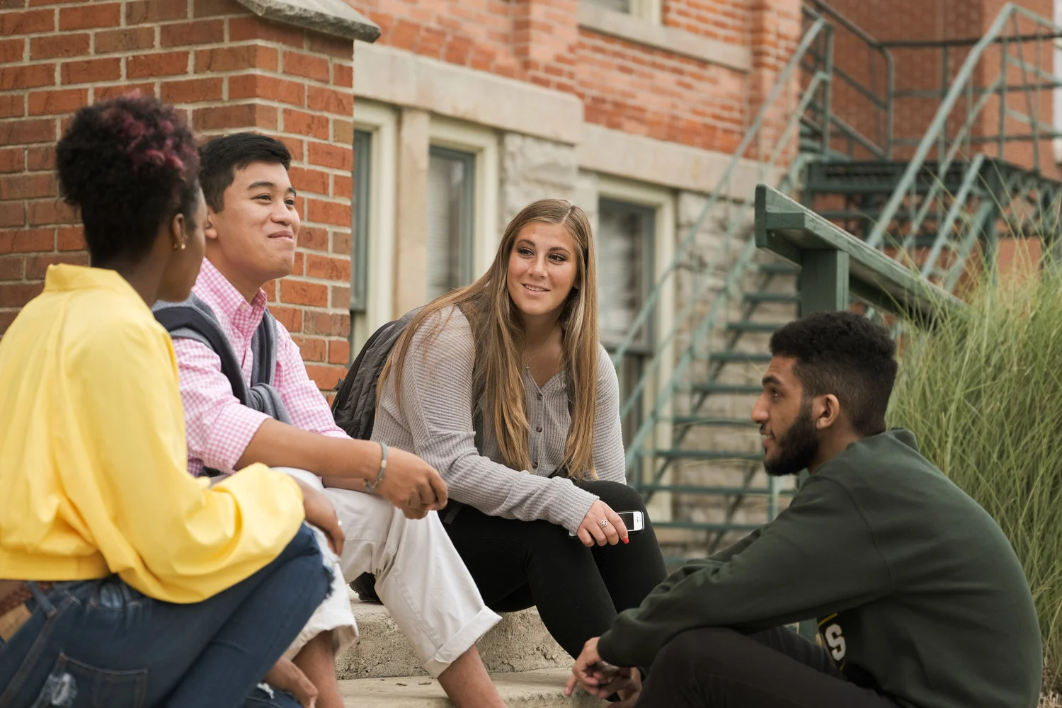 Four young people sitting on a sidewalk near a brick building, engaging in a conversation.