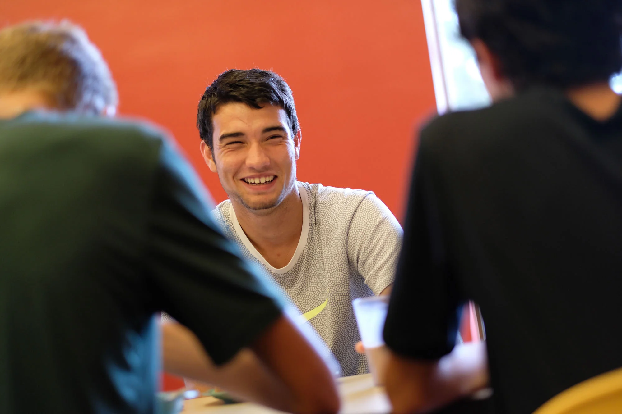 A young man with short dark hair smiling and laughing while sitting at a table with two other people, in front of a red wall.