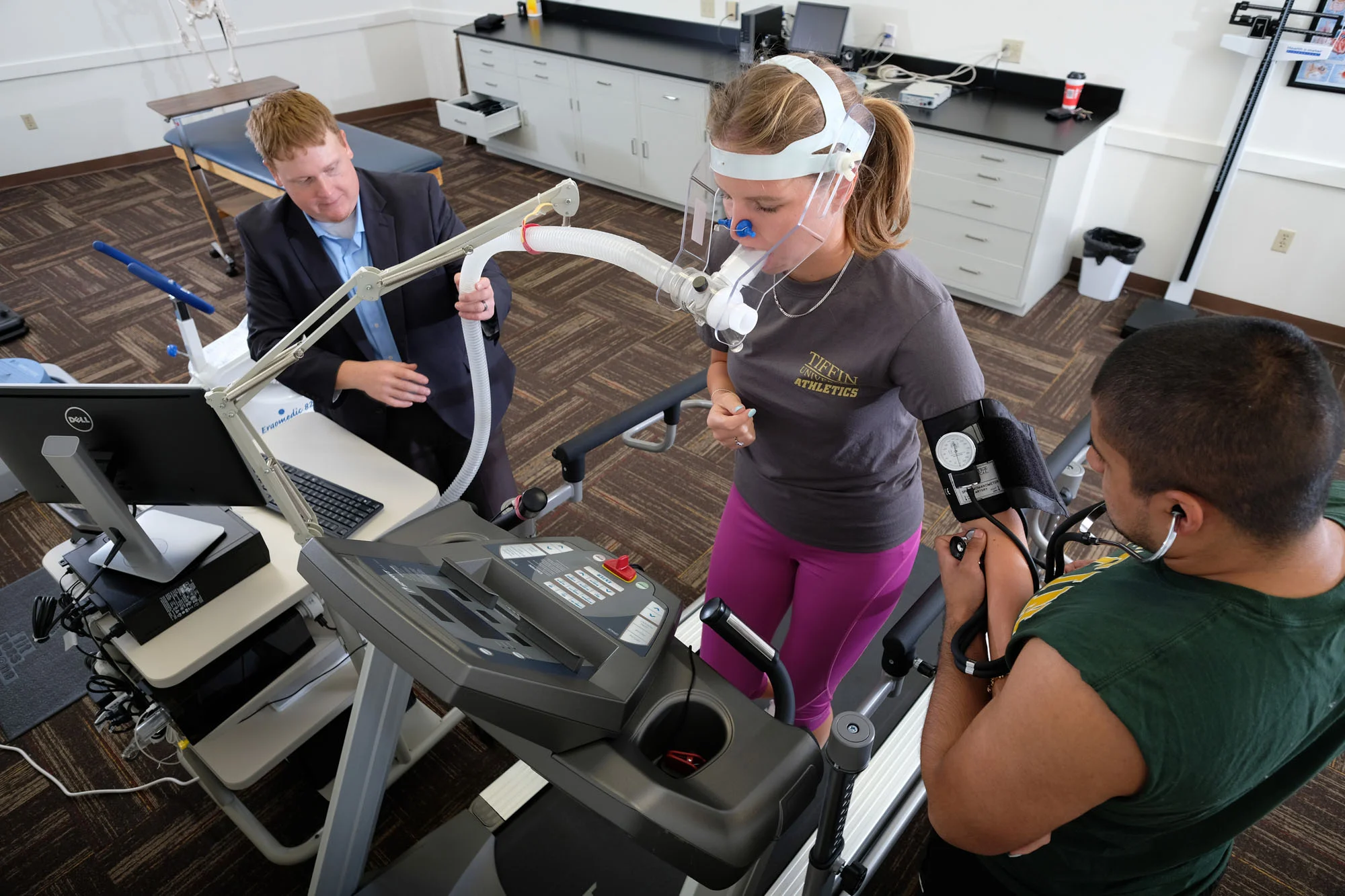 A woman wearing a mask and headset is walking on a treadmill with medical sensors attached to her arm, while two men in medical attire monitor her health in a clinical setting.