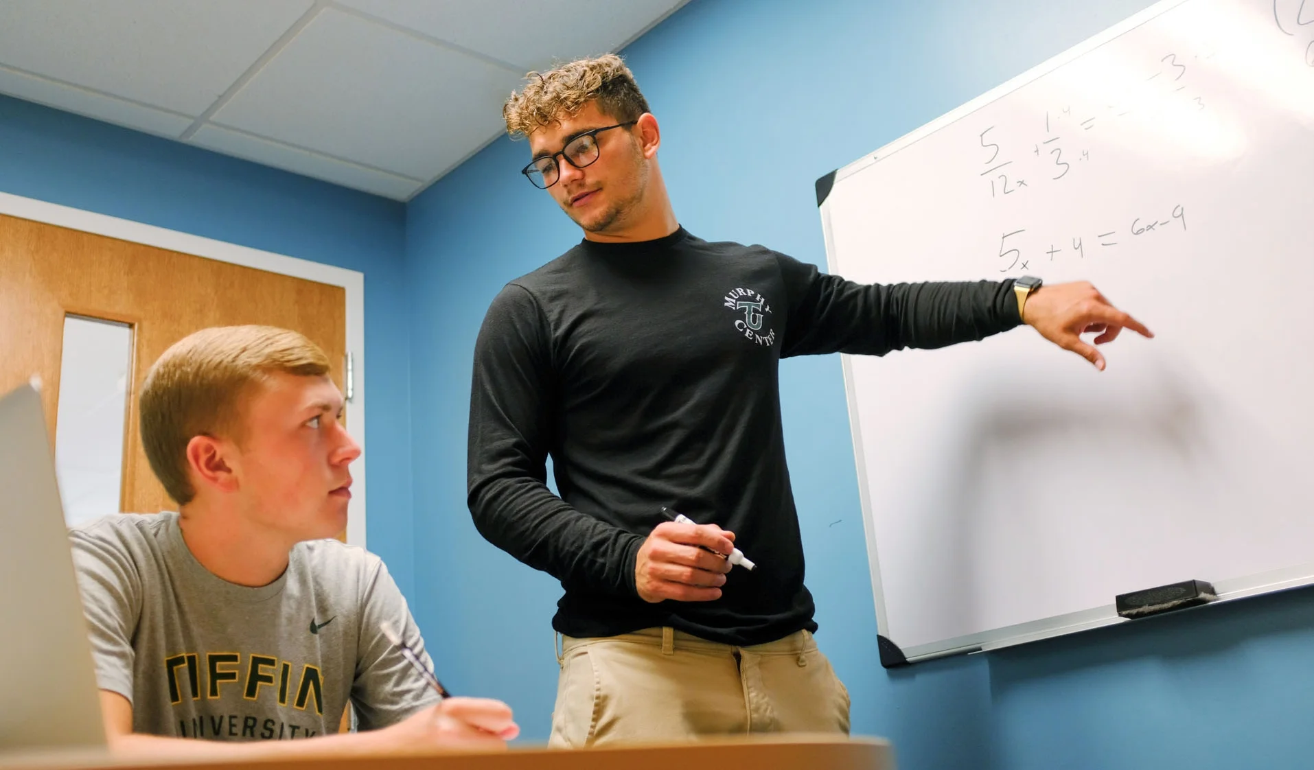 A teacher wearing glasses and a black long-sleeve shirt is pointing at a whiteboard with mathematical equations, while a student wearing a gray T-shirt sits at a desk, listening attentively.