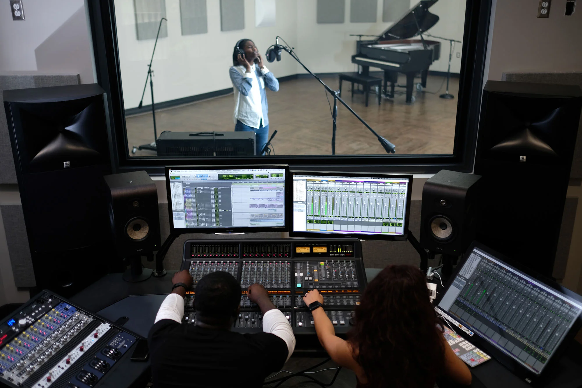 Recording studio control room with two sound engineers working at mixing consoles, two computer monitors displaying audio tracks, large speakers, and a soundproof glass window with a young woman singing into a microphone, wearing headphones, in the v
