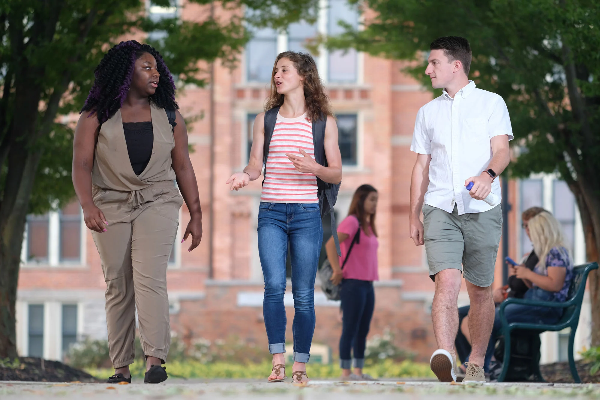 Three young adults walking and talking on a college campus with brick buildings and trees in the background, two women and one man, some people sitting and using their phones in the background.
