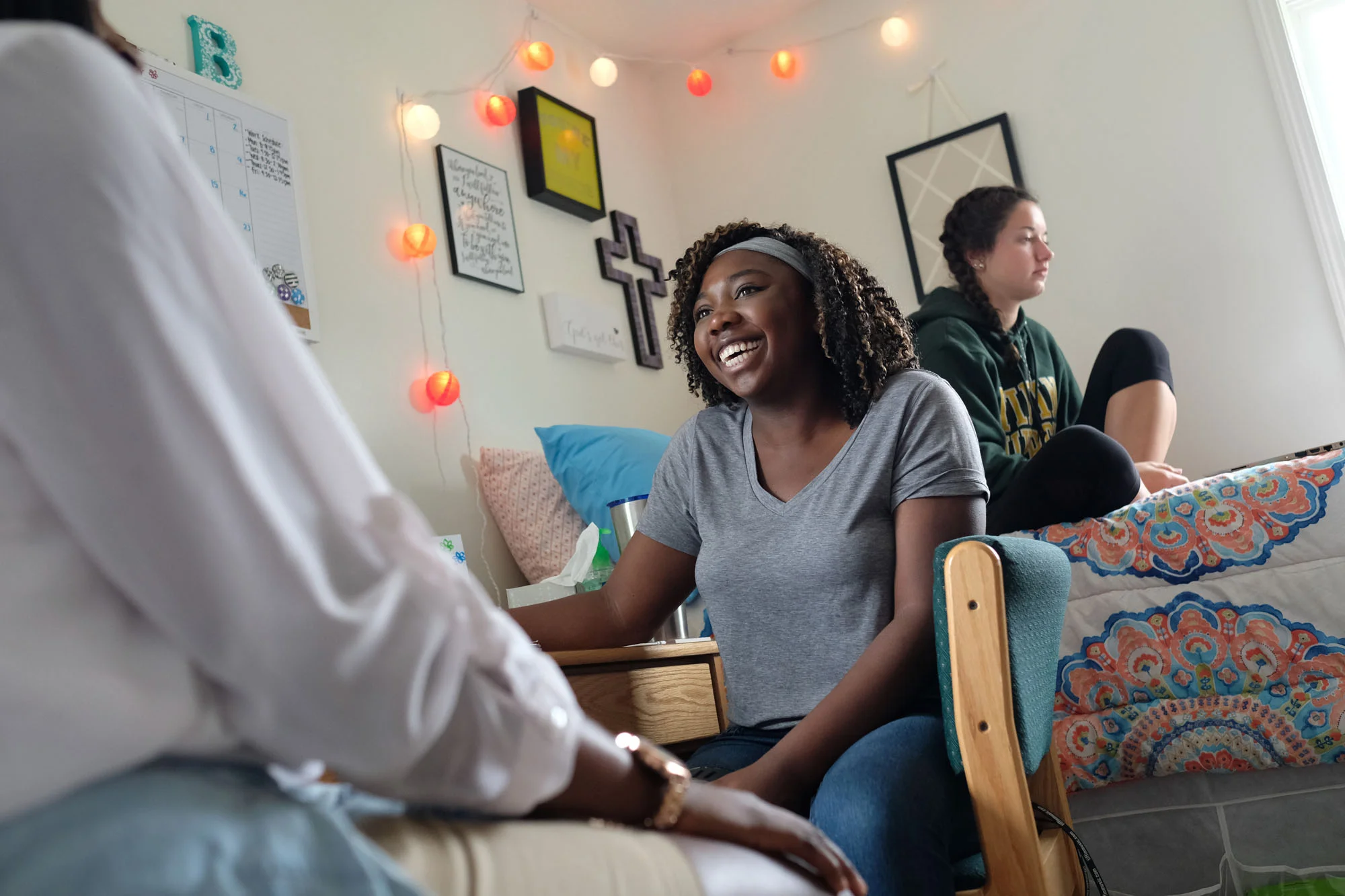 A young woman smiling and talking, sitting in a dorm room with wall decorations, string lights, and a bed nearby.