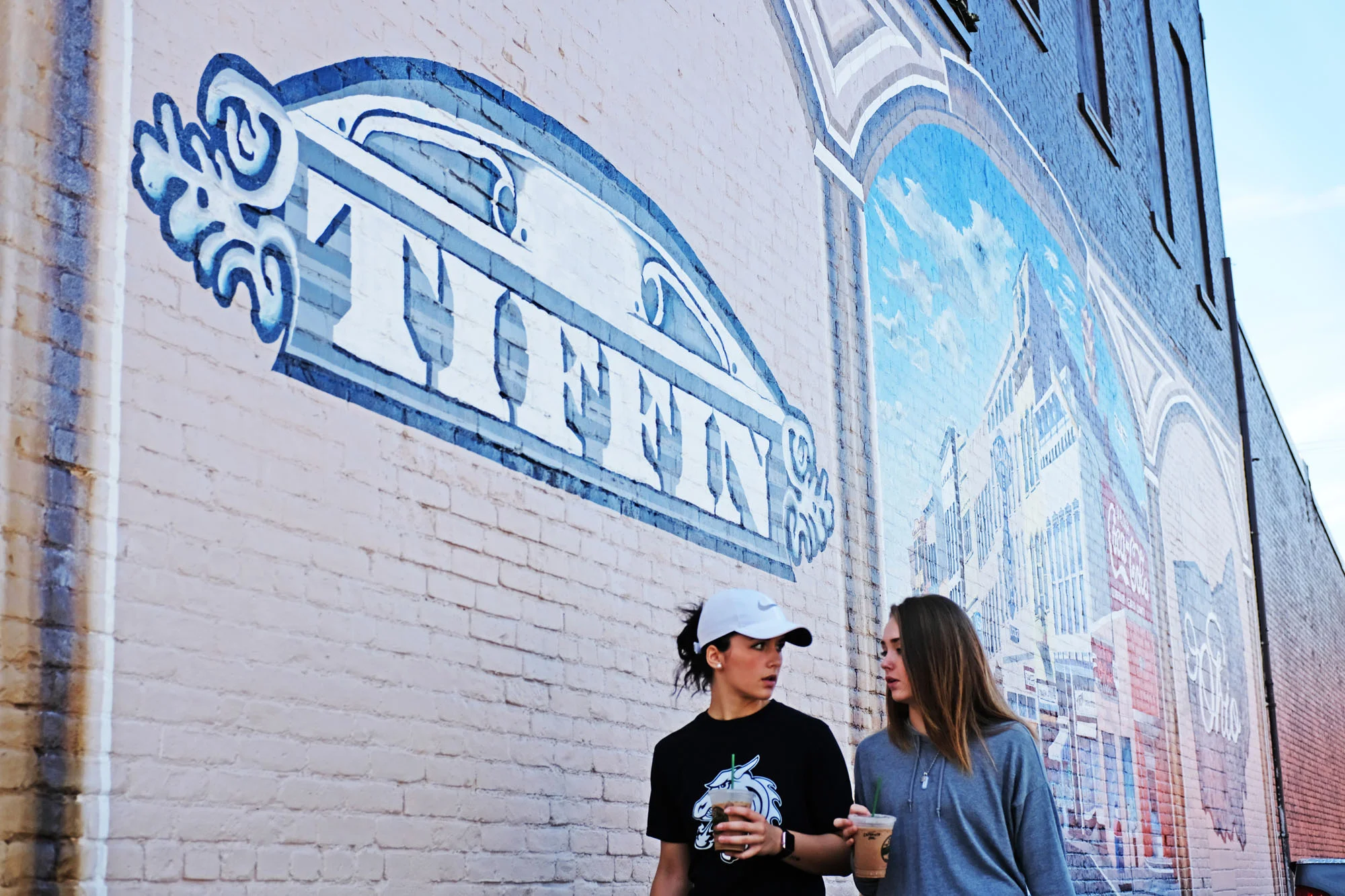 Two young women walk together in front of a large mural on a brick wall, holding iced drinks and engaging in conversation.