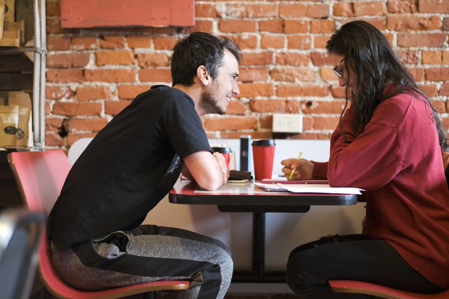 Two young adults sitting opposite each other at a table in a cafe, smiling and engaged in conversation, with coffee cups and notebooks on the table.