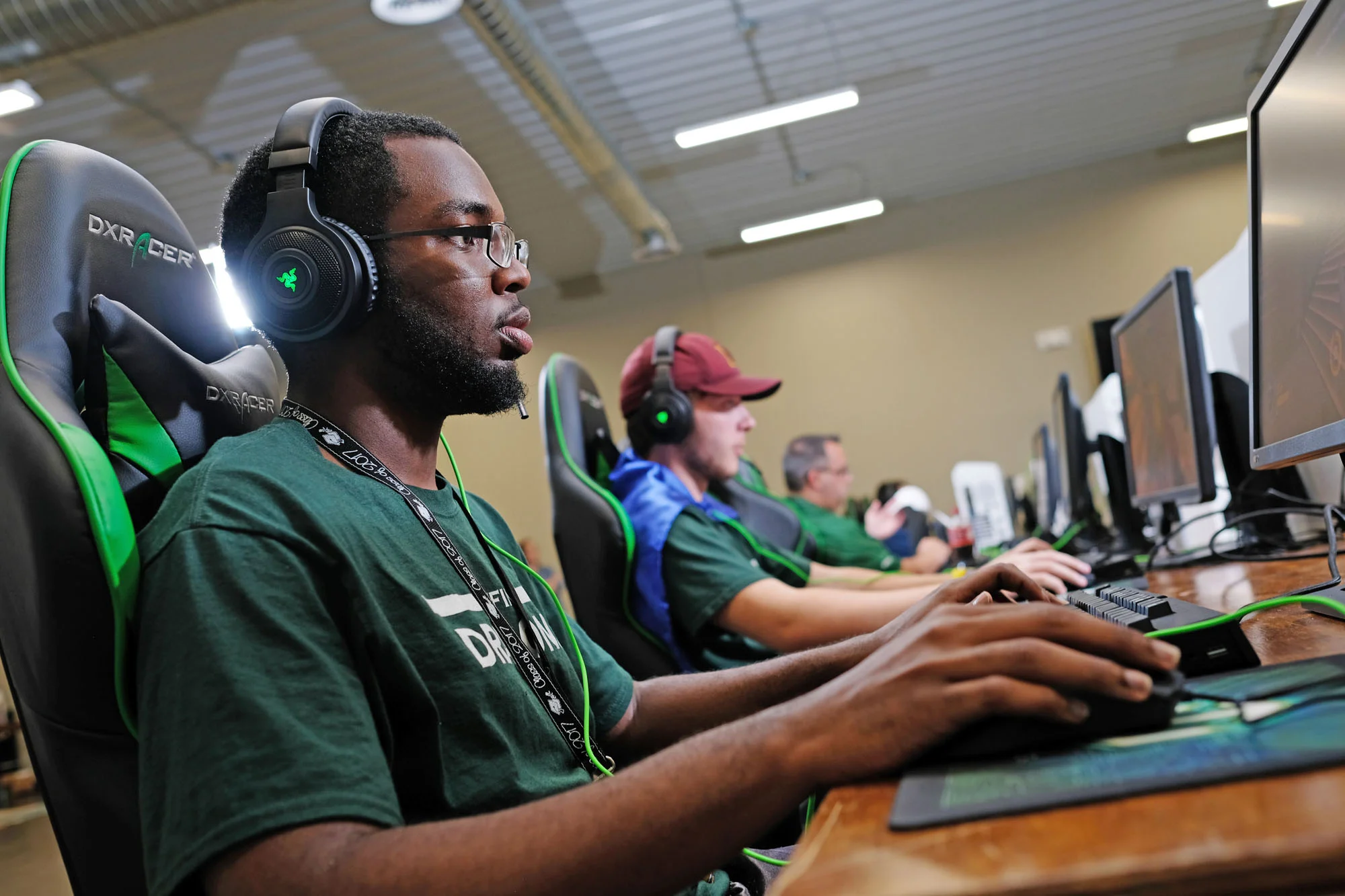 Young man with glasses using a computer at a gaming event, wearing headphones, seated in a gaming chair, with other gamers in the background.