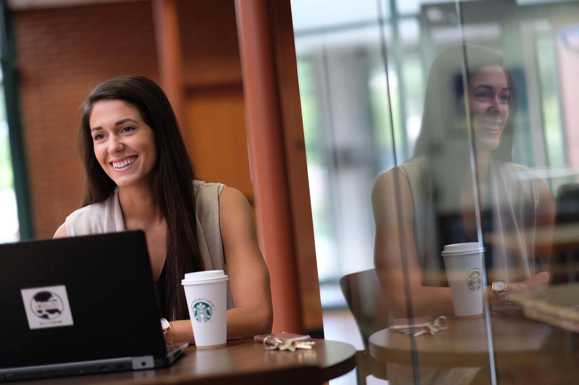 A young woman with long dark hair working on a laptop at a coffee shop, smiling, with a Starbucks cup on the table, and its reflection visible on the glass partition.