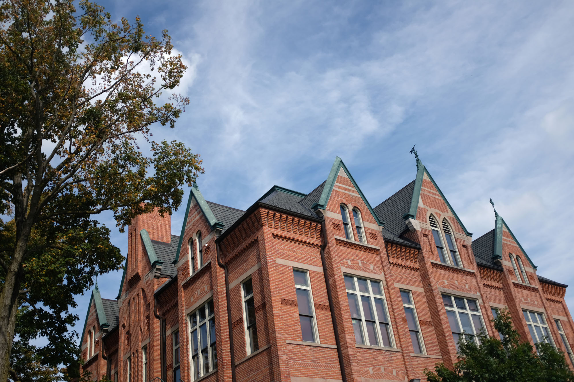 A large red brick building with tall Gothic-style windows and steep roof gables, surrounded by green trees and a blue sky with some clouds.
