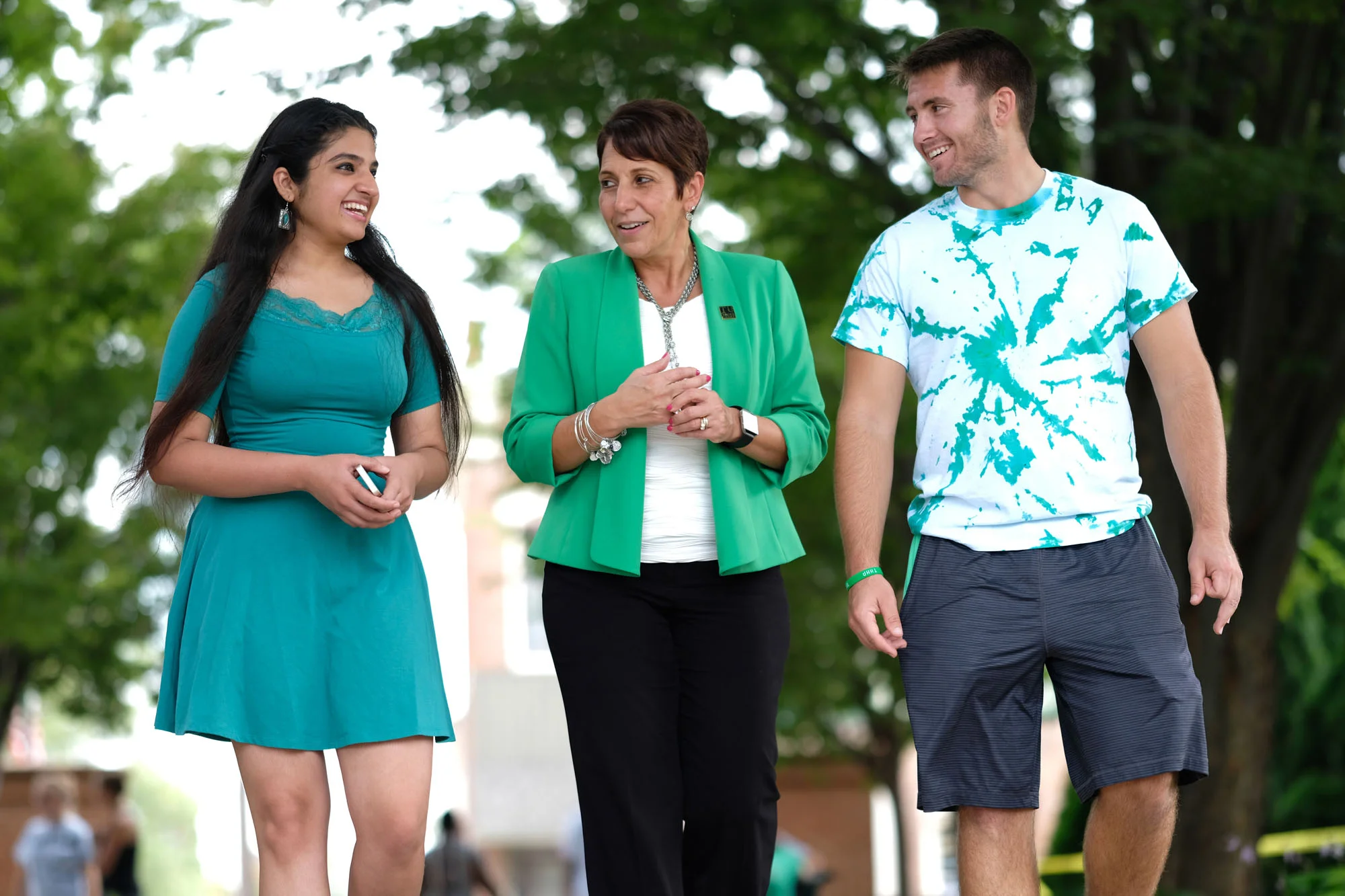 Three people, a young woman, an older woman, and a young man, walking and talking outdoors in a park with green trees in the background.