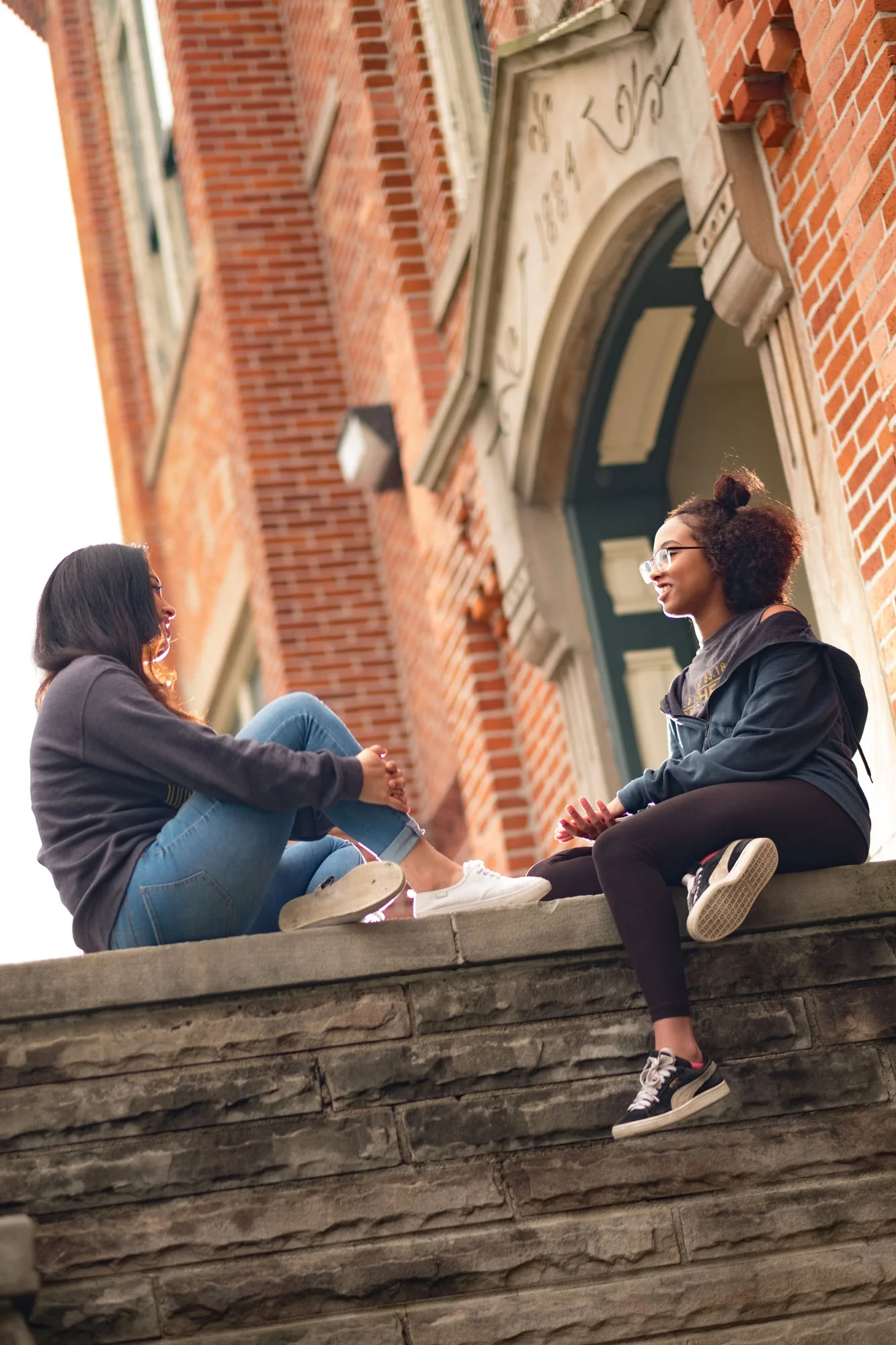 Two young women sitting on a stone ledge outside a brick building, talking and smiling at each other.