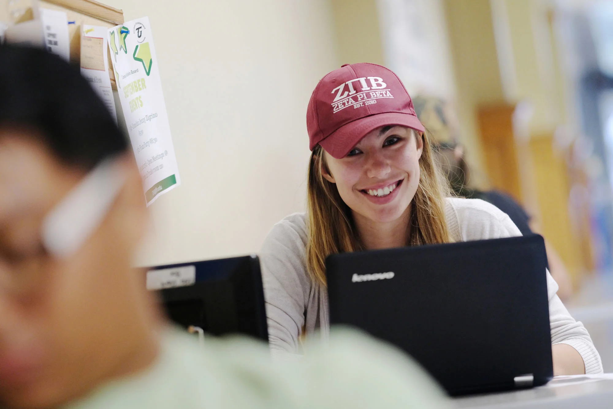 A young woman with long blonde hair wearing a red baseball cap and a white shirt, smiling at a laptop computer in a room with light-colored walls and posters.