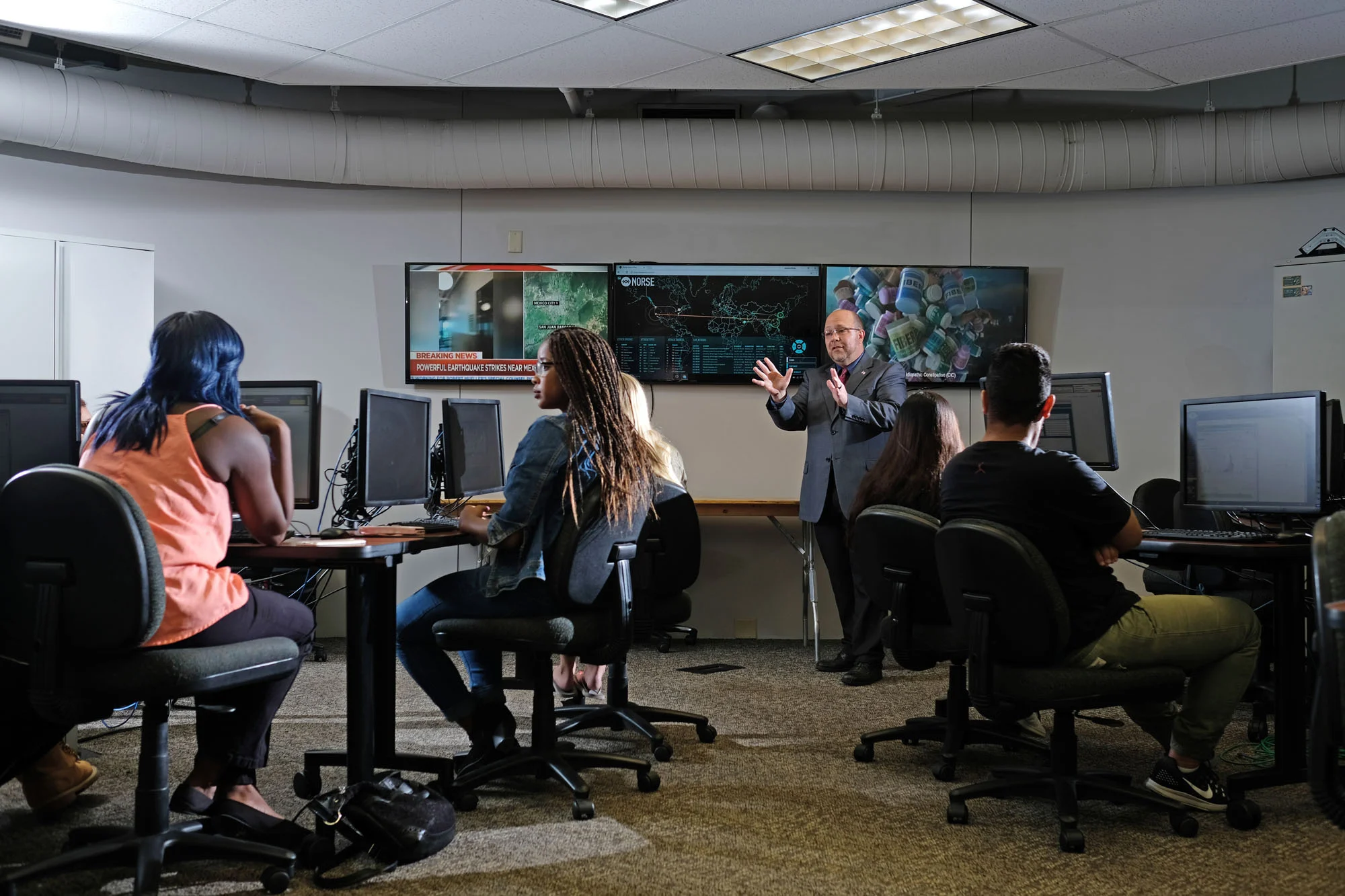 A classroom or training room with students sitting at desks with computers, listening to an instructor who is standing in front of large screens displaying maps, news, and pills.