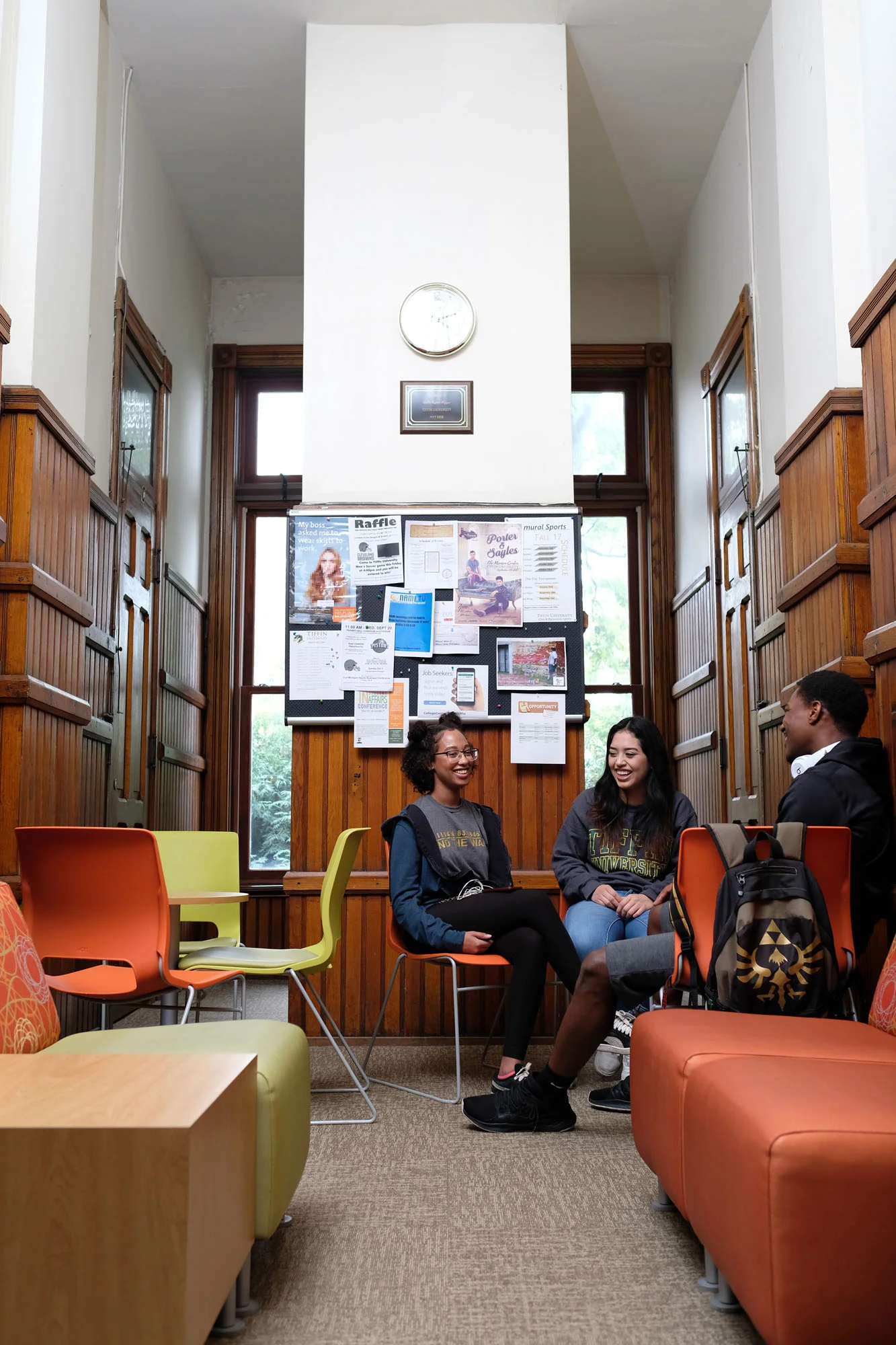 Three students sitting on chairs and talking in a cozy, wood-paneled common room with bulletin boards on the wall and large windows.