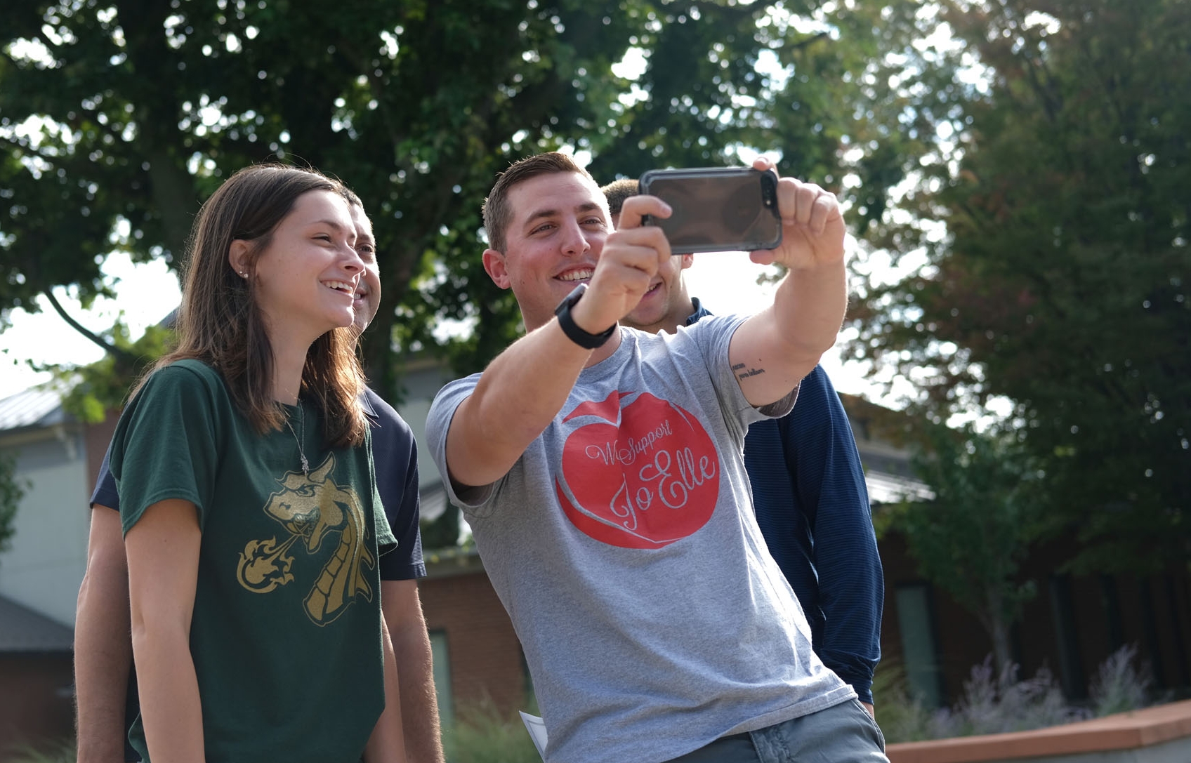 A group of young people taking a selfie outdoors on a sunny day, with trees in the background.