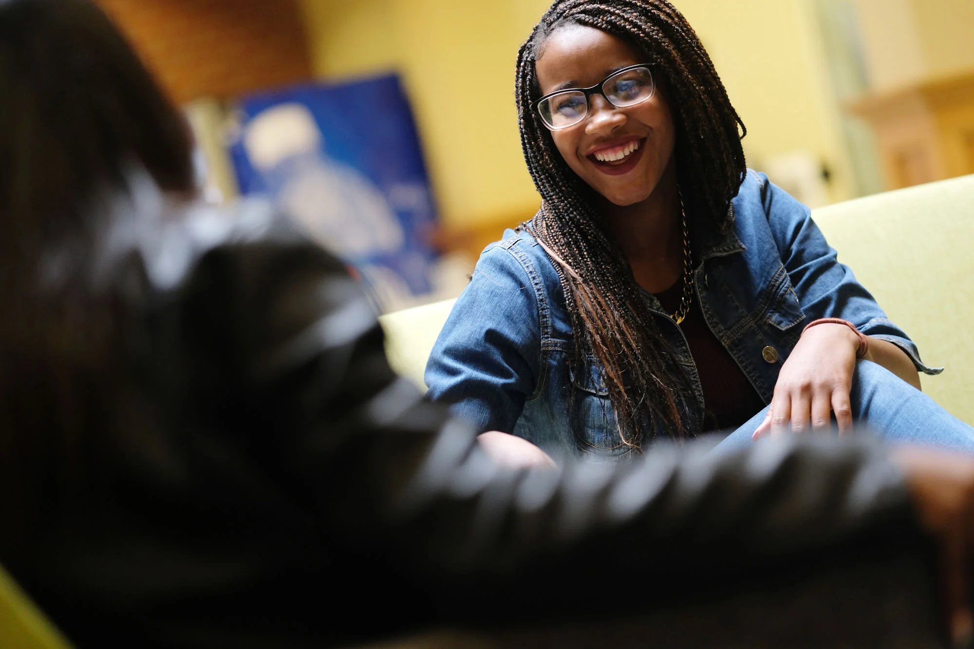 A woman with glasses and curly braided hair, smiling and talking, wearing a denim jacket, sitting on a yellow chair in a cozy indoor setting.