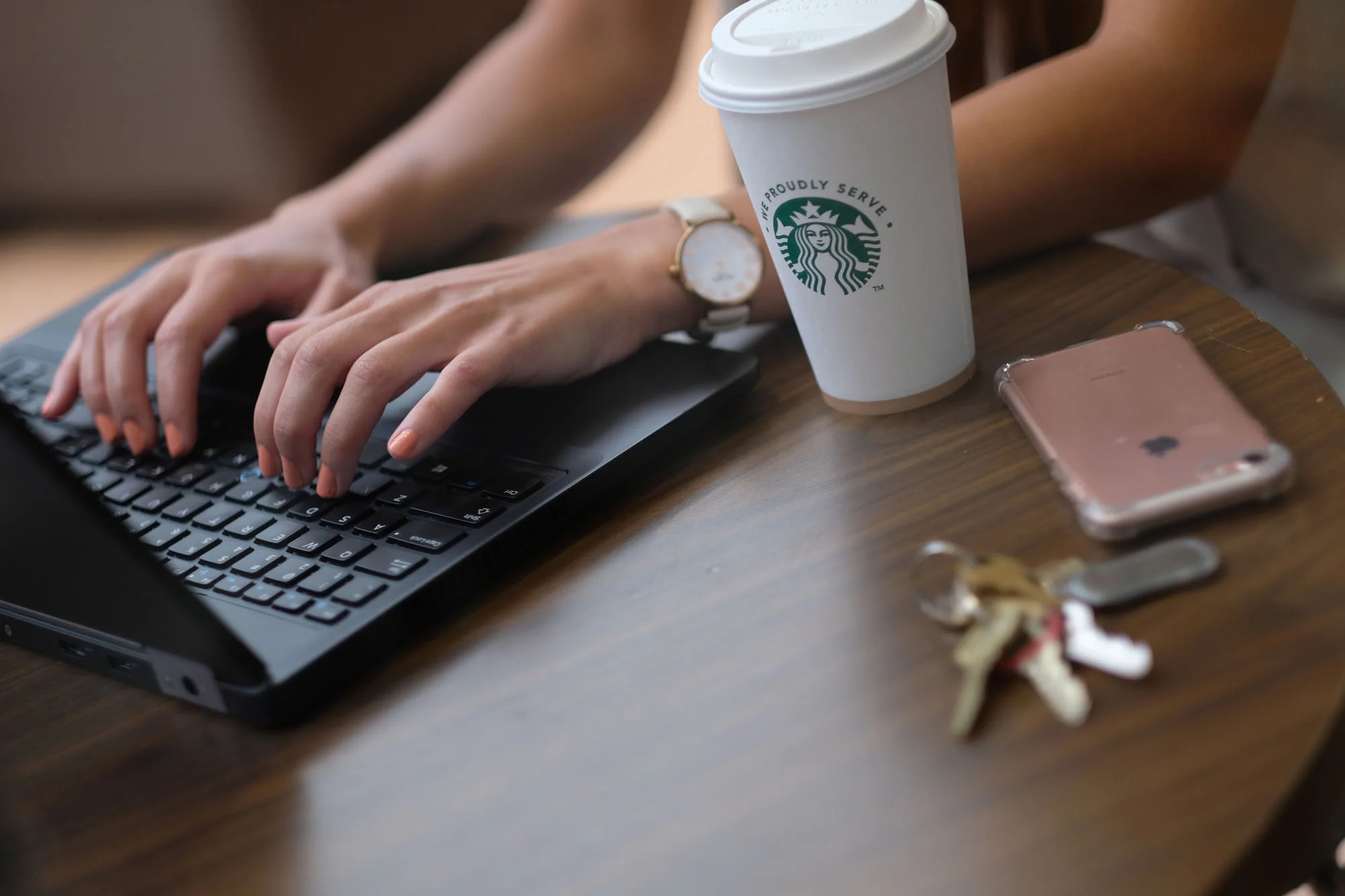 Person typing on a black laptop at a wooden table with a Starbucks coffee cup, a pink iPhone, a set of keys, and a small figurine.