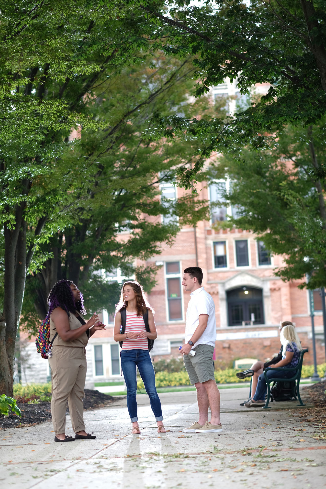 Three young people standing and talking in a park with trees and a brick building in the background. Two women sitting on a park bench nearby.