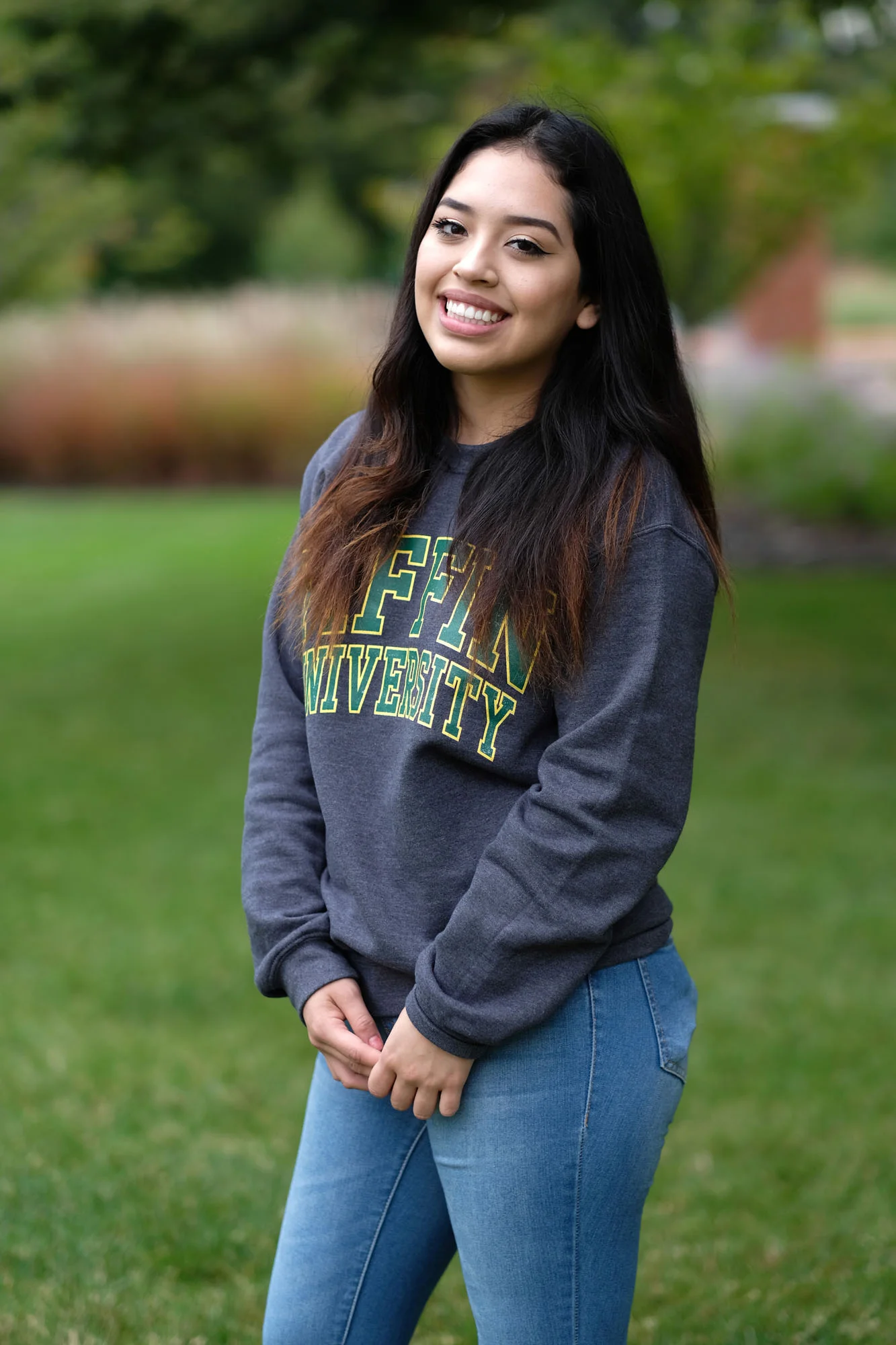 A young woman outdoors with green grass and trees in the background, smiling and wearing a gray sweatshirt with campus lettering and blue jeans.