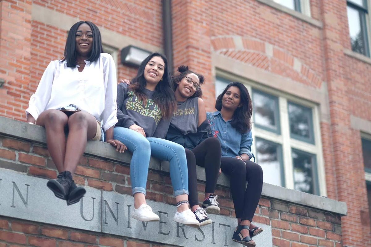 Four young women sitting on a brick ledge in front of a brick building, smiling at the camera.