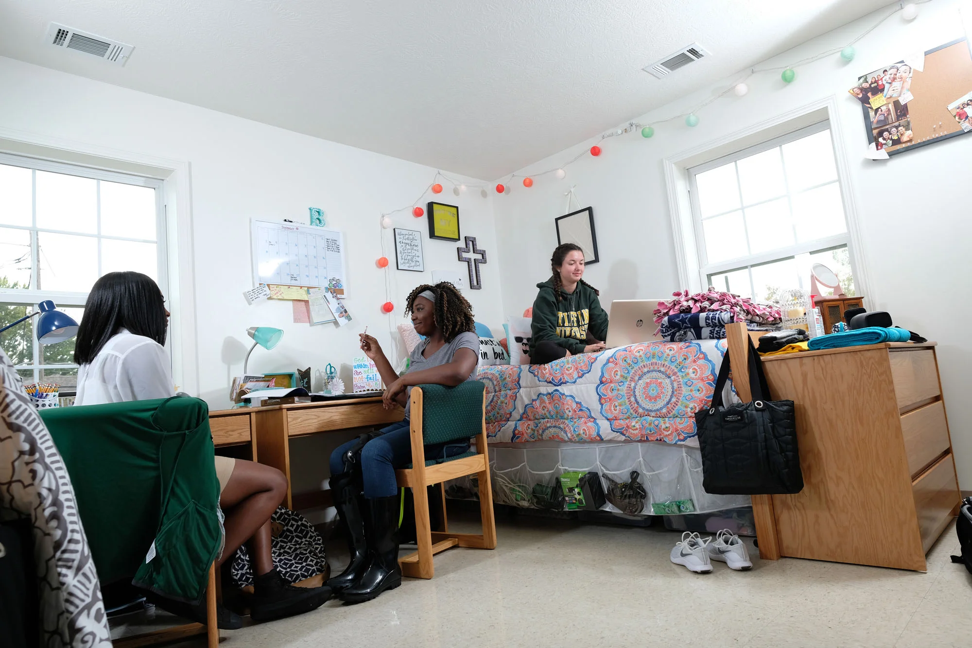 Three young women in a dorm room with large windows, engaged in conversation and studying, with colorful decorations and personal items around.